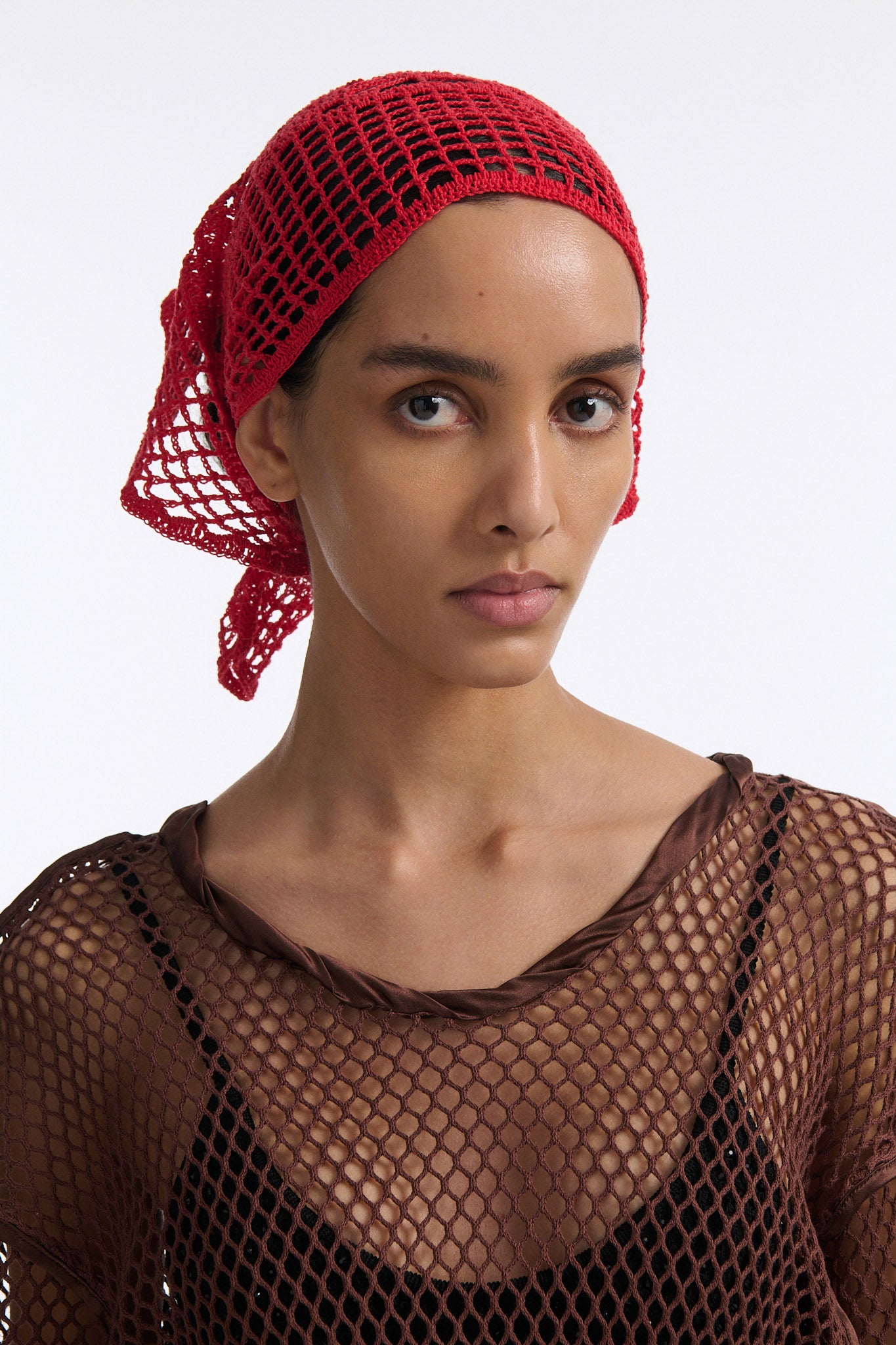 Female model with slicked back black hair in a bun wears brown see-through mesh long sleeve top and bright red crochet bandana tied around her forehead.