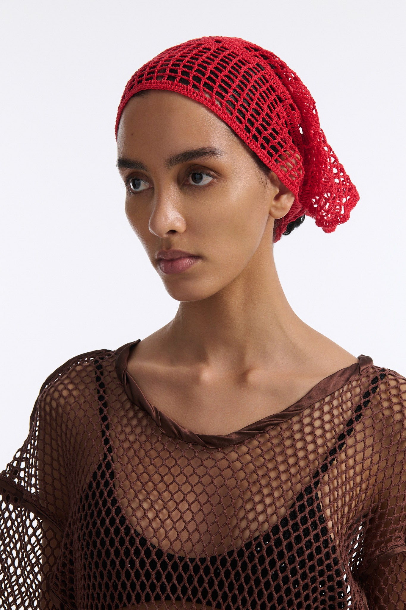 Female model with slicked back black hair in a bun wears brown see-through mesh long sleeve top and bright red crochet bandana tied around her forehead.