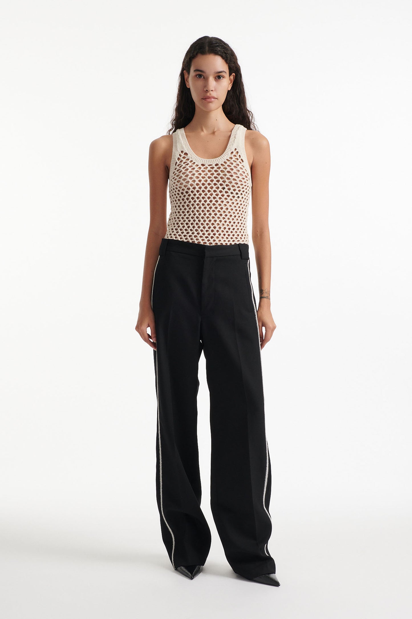 Female model wears beige see-through mesh tank top, wide black trousers with white stitched stripes, and black high heel boots on white background.