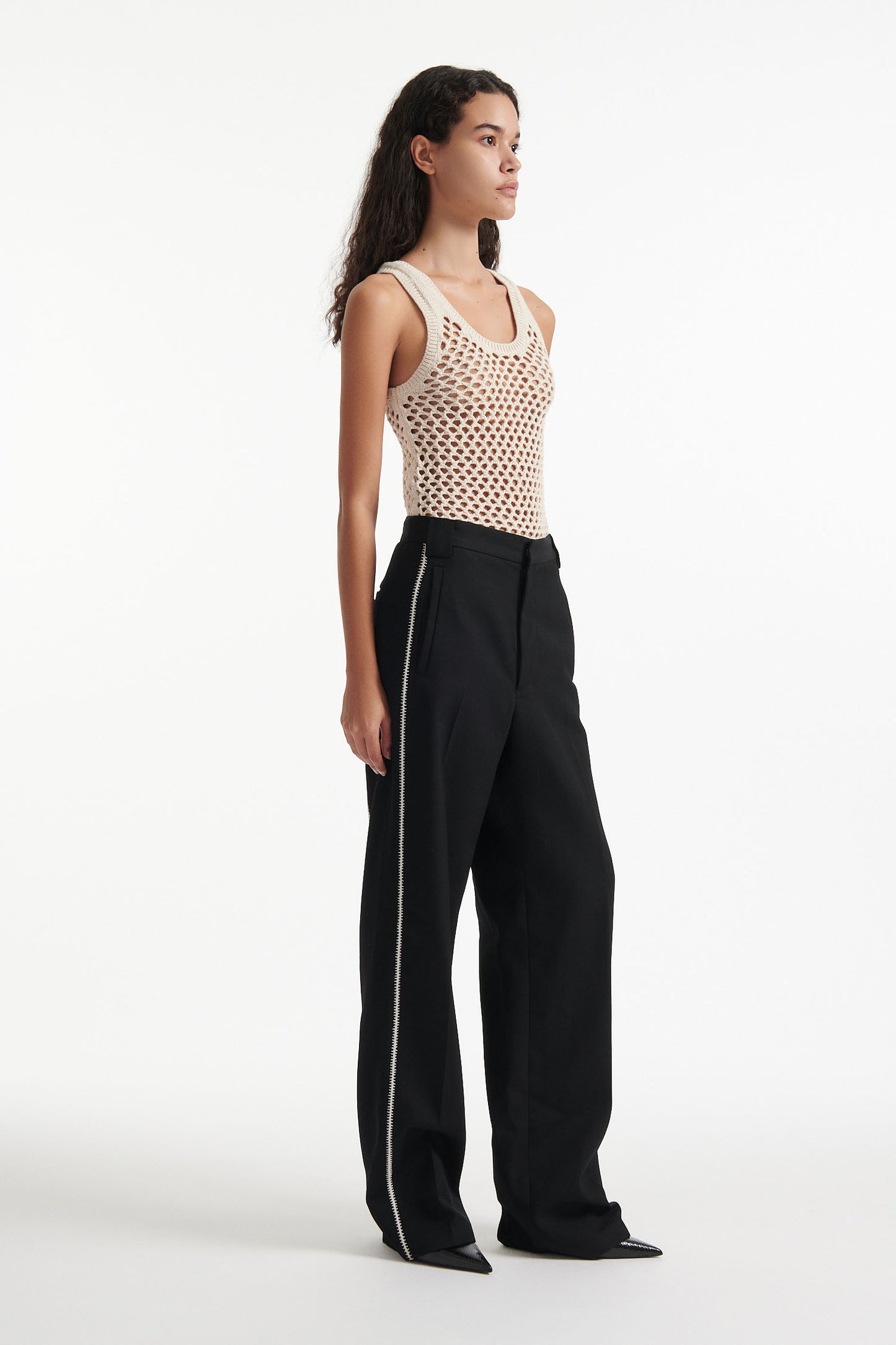 Female model wears beige see-through mesh tank top, wide black trousers with white stitched stripes, and black high heel boots on white background.