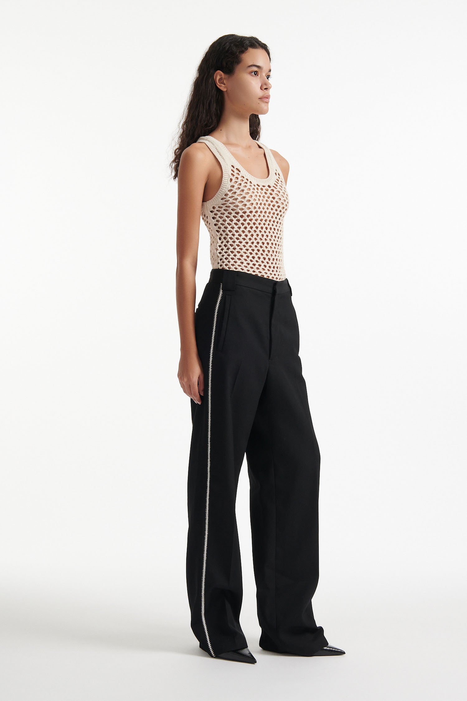 Female model wears beige see-through mesh tank top, wide black trousers with white stitched stripes, and black high heel boots on white background.