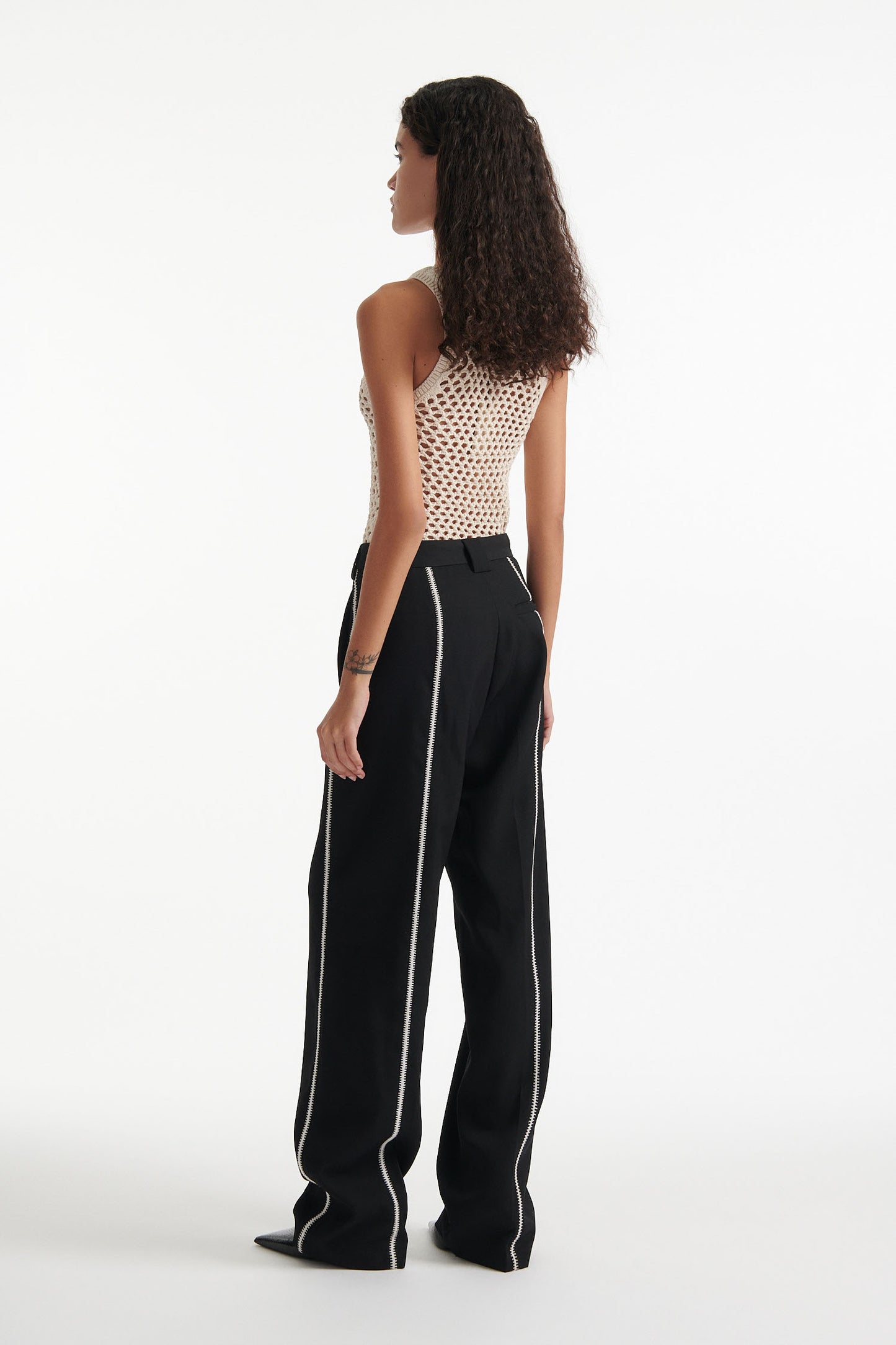 Female model wears beige see-through mesh tank top, wide black trousers with white stitched stripes, and black high heel boots on white background.