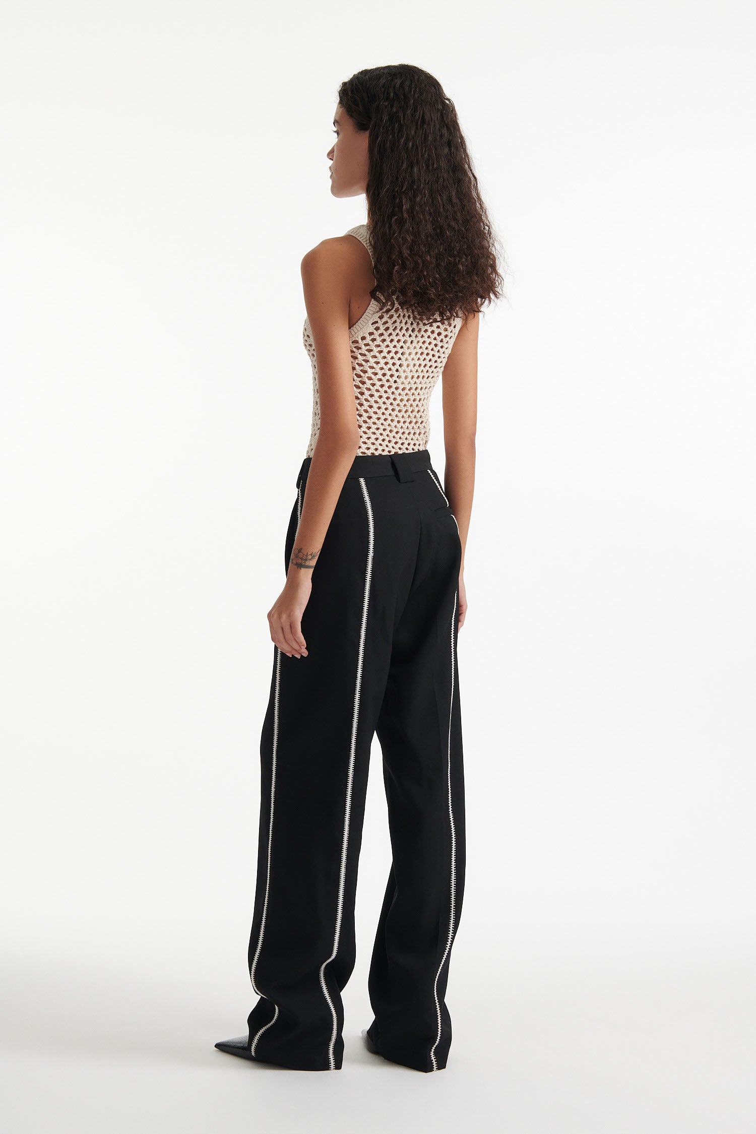 Female model wears beige see-through mesh tank top, wide black trousers with white stitched stripes, and black high heel boots on white background.