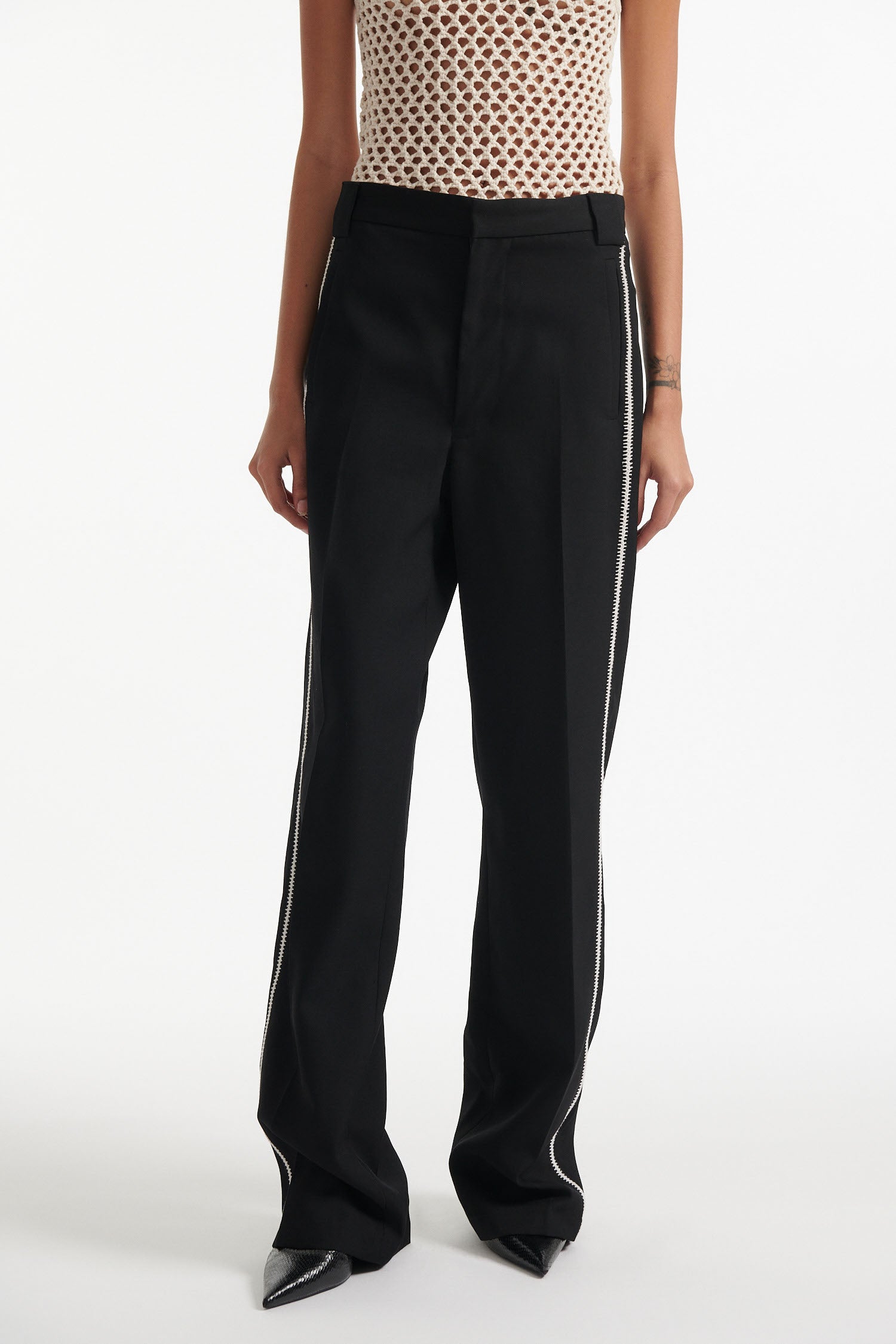 Female model wears beige see-through mesh tank top, wide black trousers with white stitched stripes, and black high heel boots on white background.