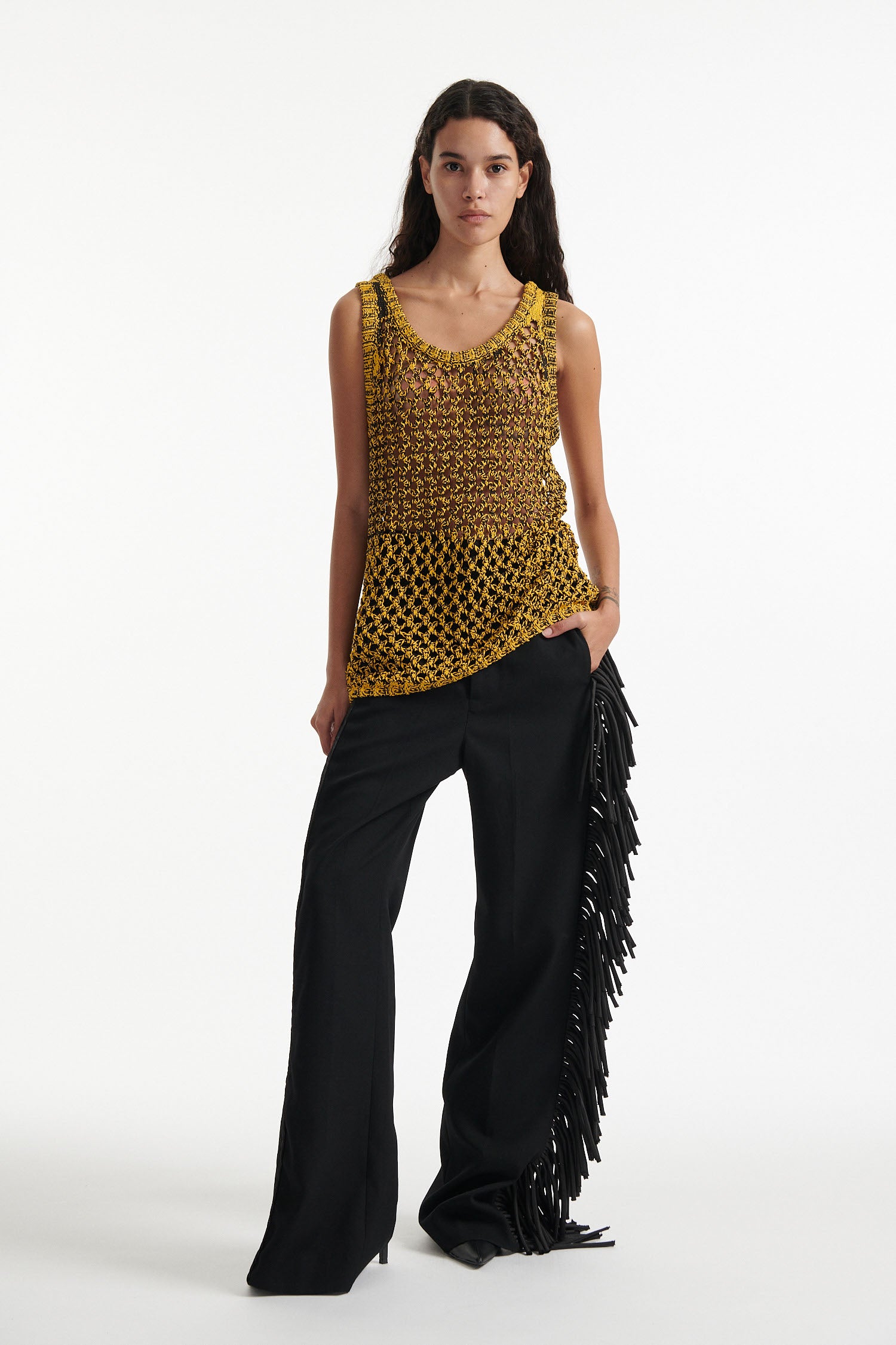 Female model wears yellow mesh tank dress, black fringe trousers, black high heel boots, and chunky silver bracelet on white background.