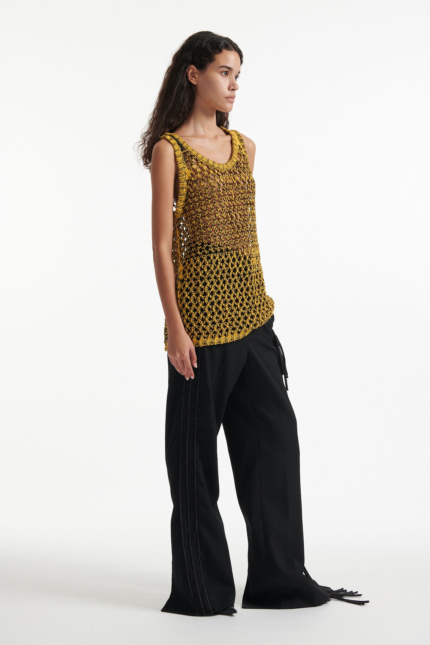 Female model wears yellow mesh tank dress, black fringe trousers, black high heel boots, and chunky silver bracelet on white background.