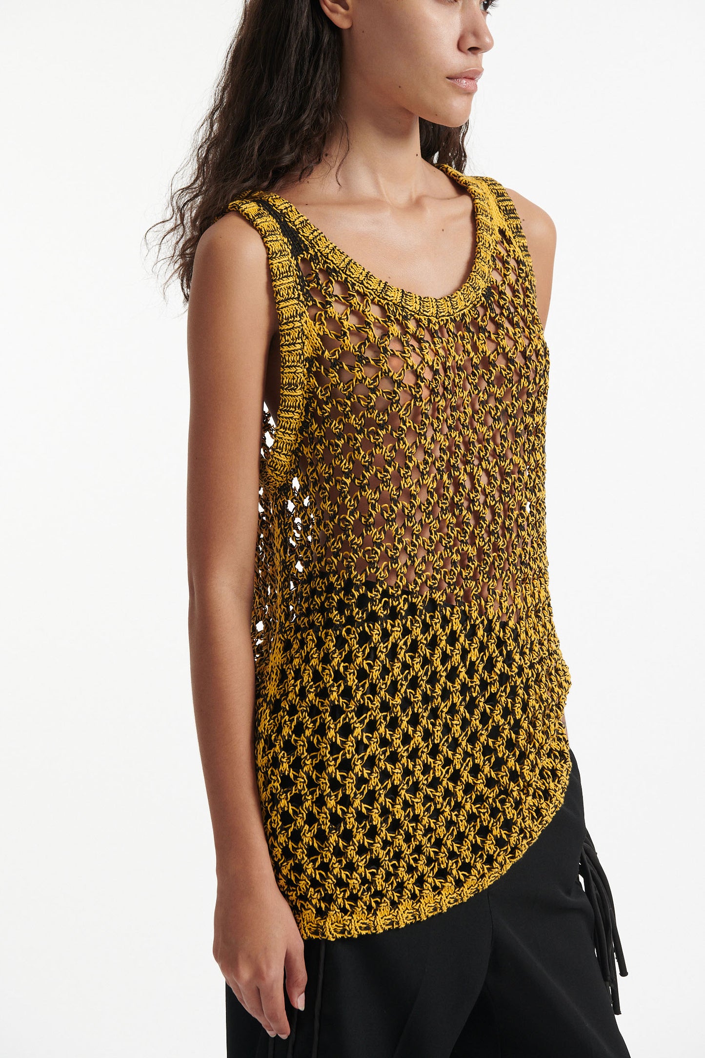 Female model wears yellow mesh tank dress, black fringe trousers, black high heel boots, and chunky silver bracelet on white background.