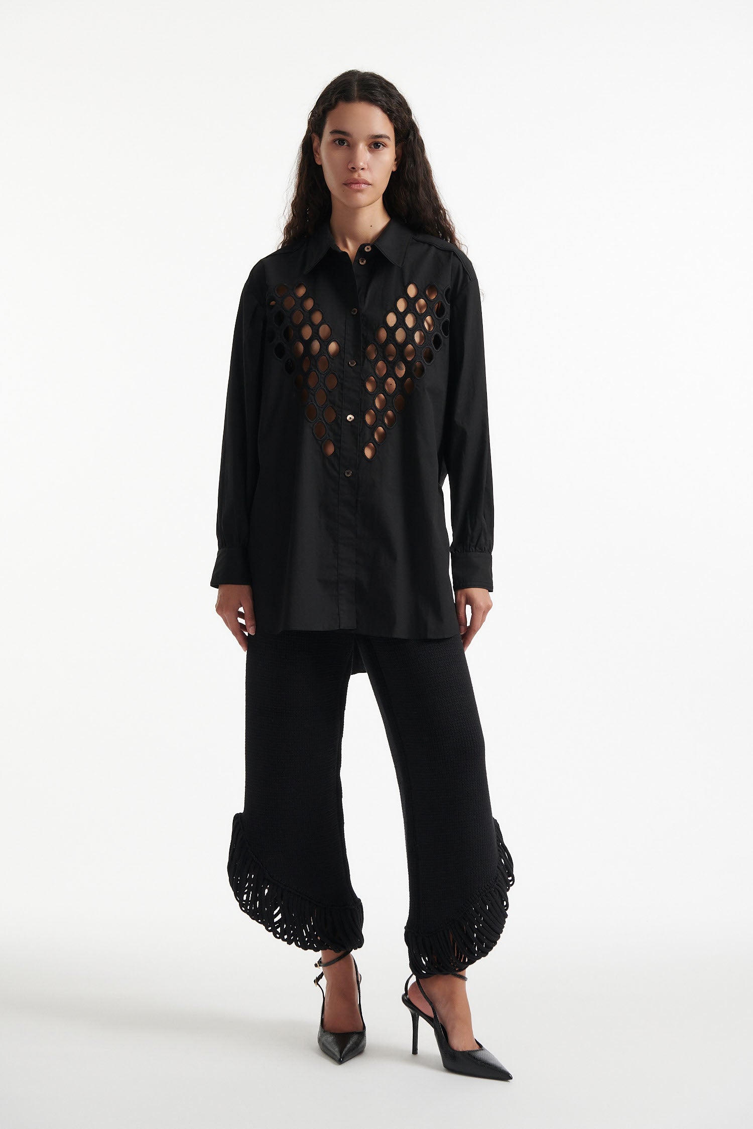 Female model wears black cotton shirt with see-through cutouts at chest and back. Model also wears black knit pants with fringe and black high heels on white background.