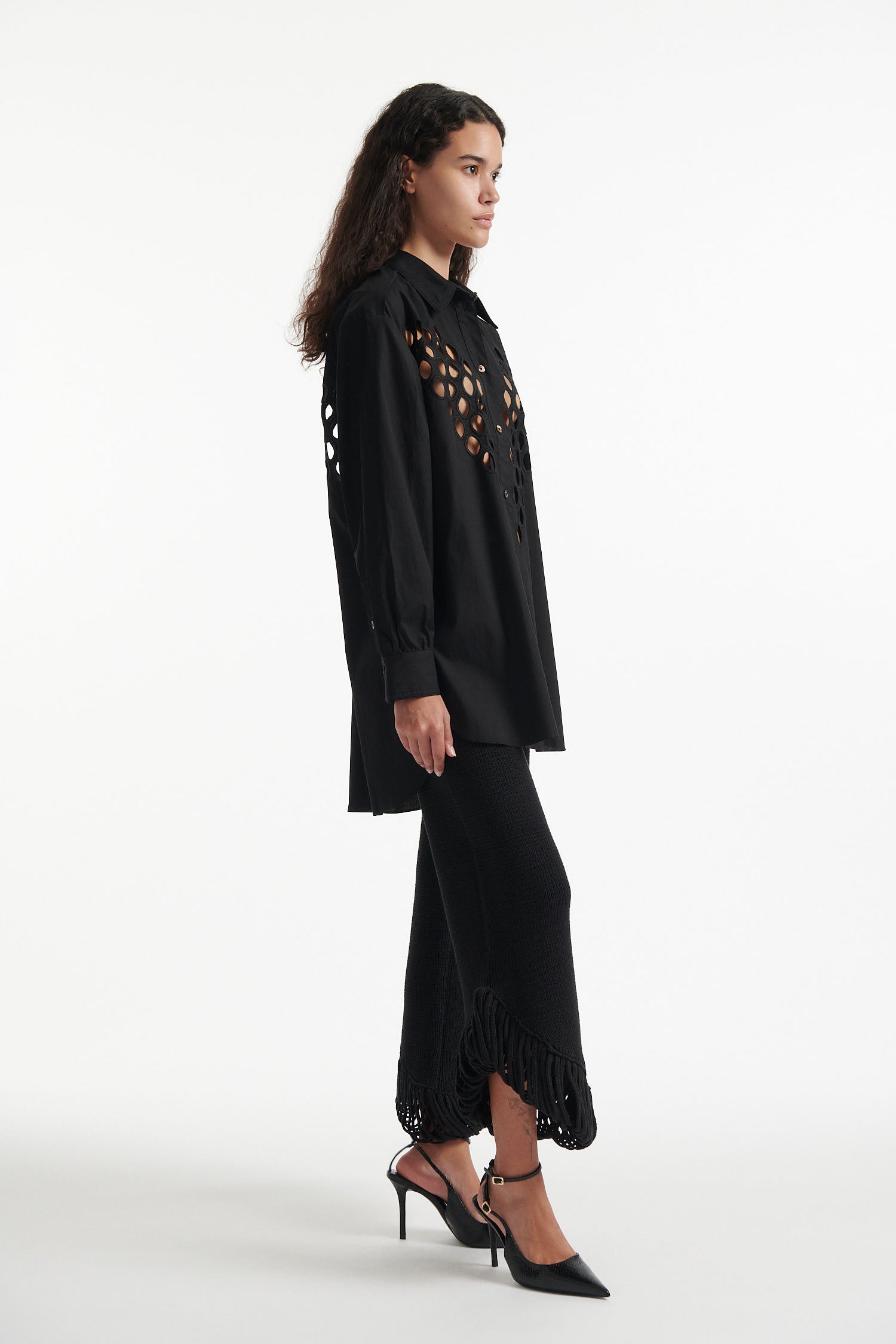 Female model wears black cotton shirt with see-through cutouts at chest and back. Model also wears black knit pants with fringe and black high heels on white background.