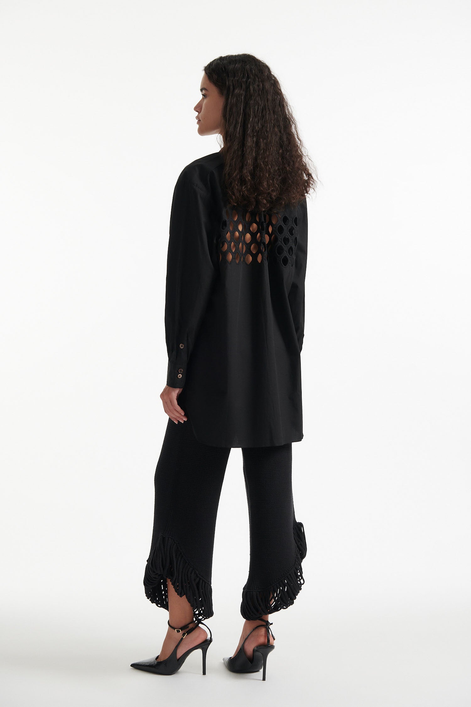 Female model wears black cotton shirt with see-through cutouts at chest and back. Model also wears black knit pants with fringe and black high heels on white background.