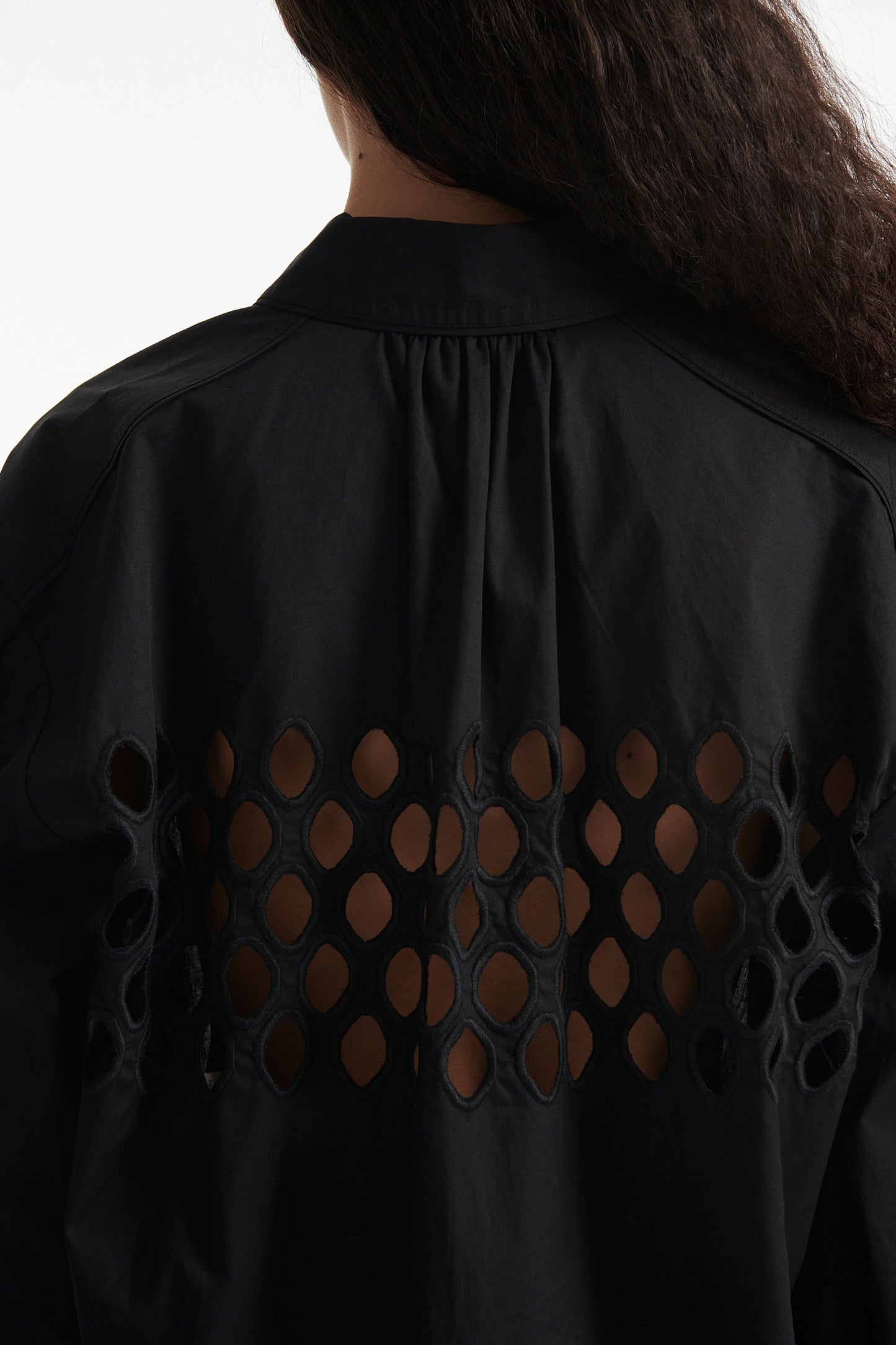 Female model wears black cotton shirt with see-through cutouts at chest and back. Model also wears black knit pants with fringe and black high heels on white background.