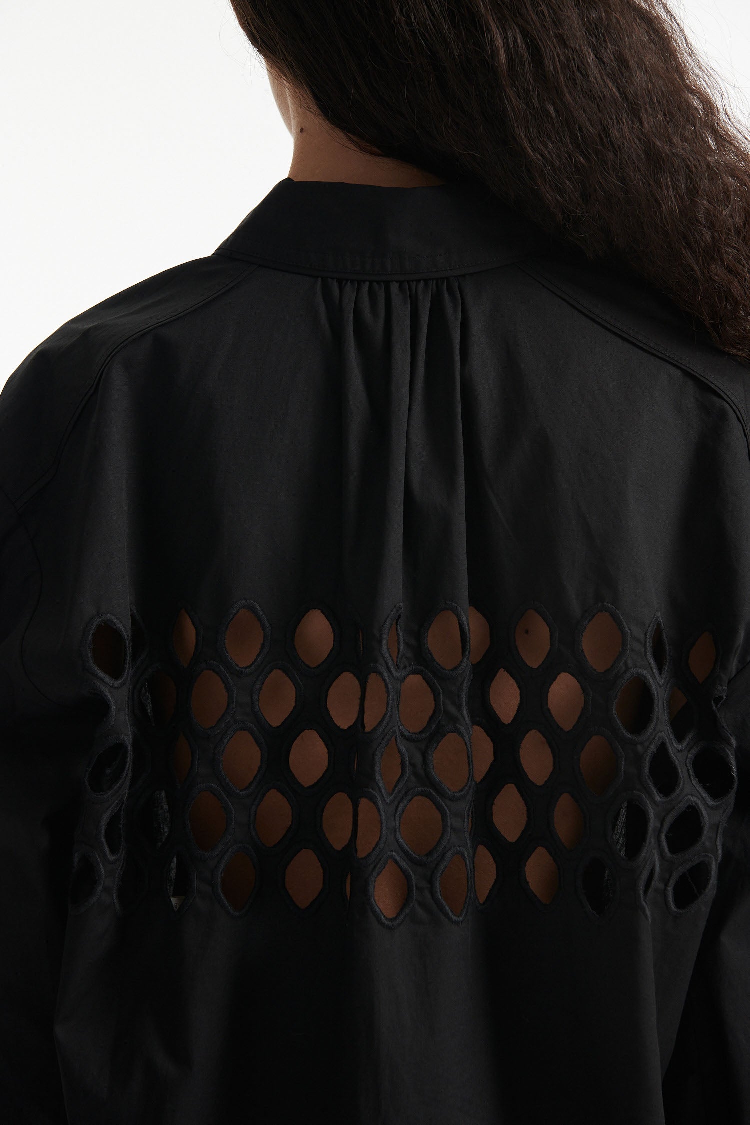 Female model wears black cotton shirt with see-through cutouts at chest and back. Model also wears black knit pants with fringe and black high heels on white background.