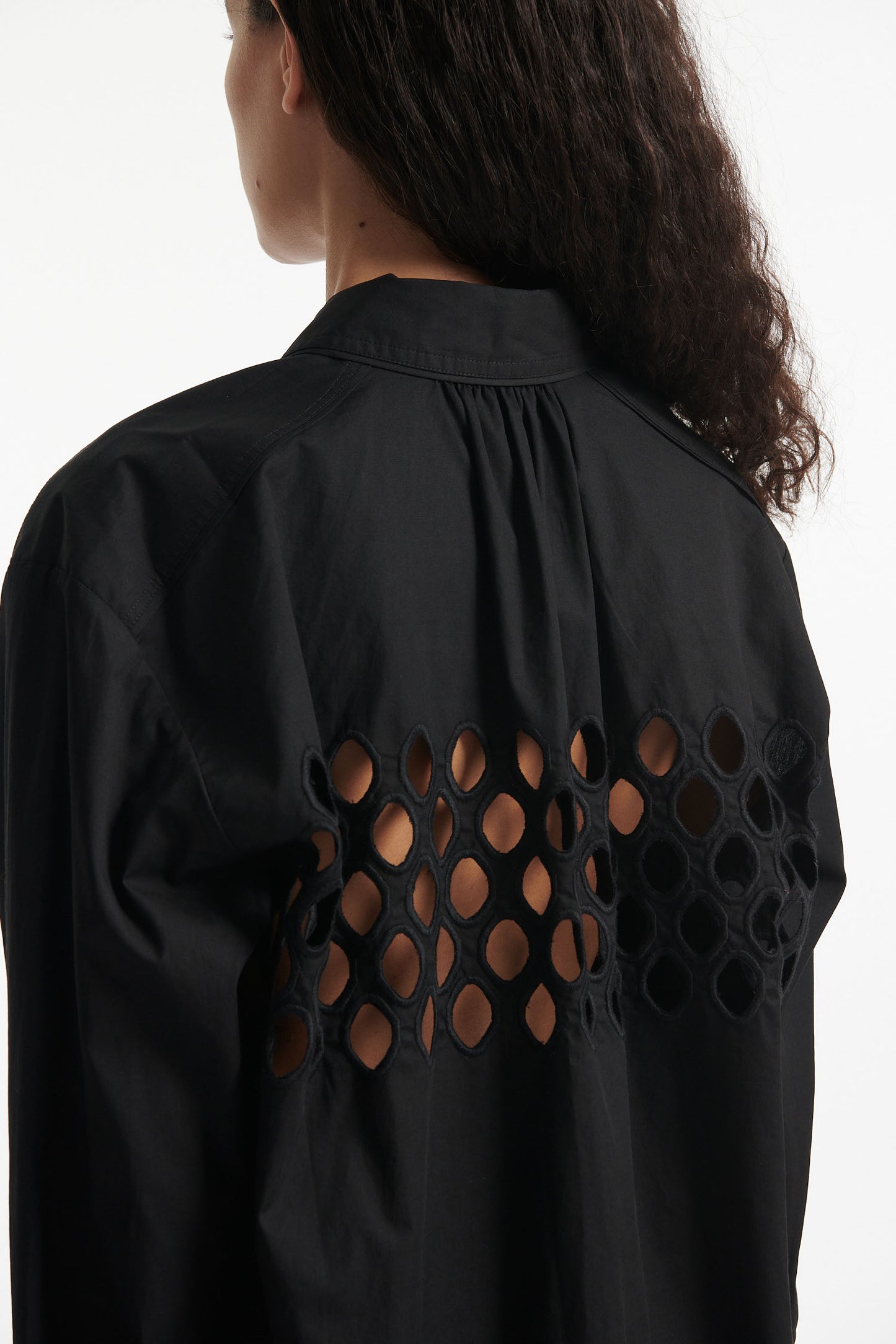 Female model wears black cotton shirt with see-through cutouts at chest and back. Model also wears black knit pants with fringe and black high heels on white background.