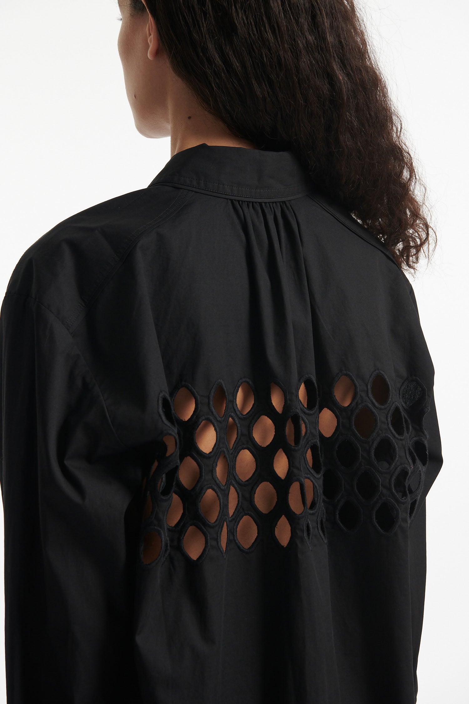 Female model wears black cotton shirt with see-through cutouts at chest and back. Model also wears black knit pants with fringe and black high heels on white background.