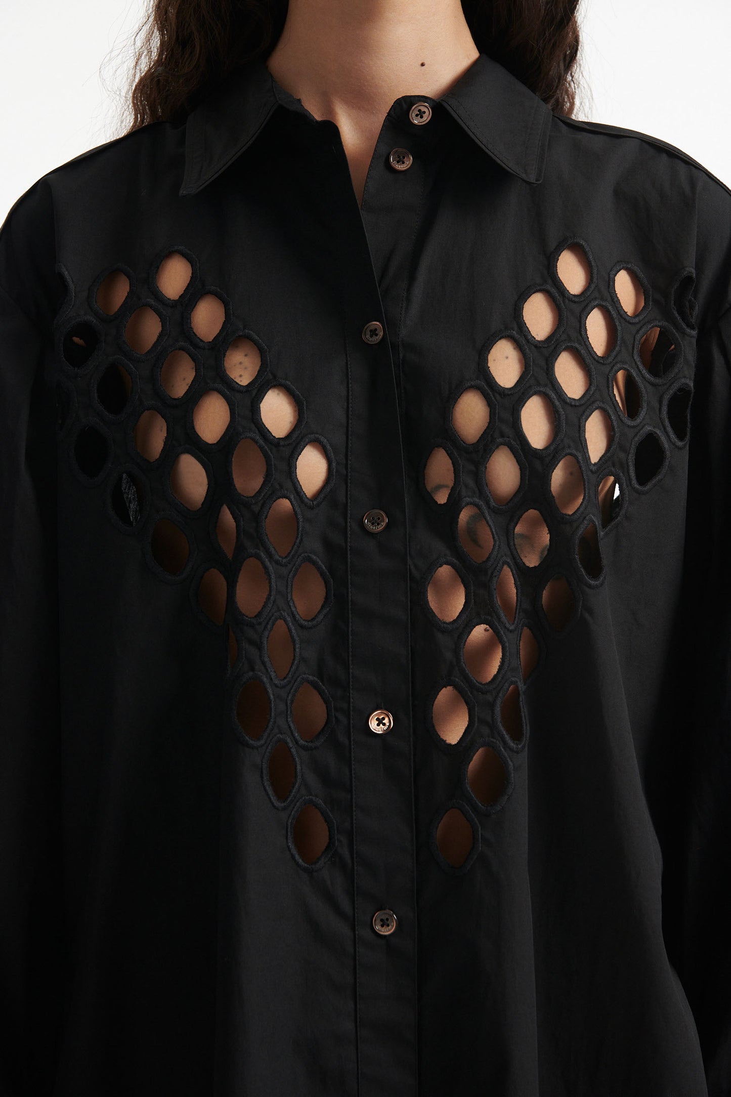 Female model wears black cotton shirt with see-through cutouts at chest and back. Model also wears black knit pants with fringe and black high heels on white background.