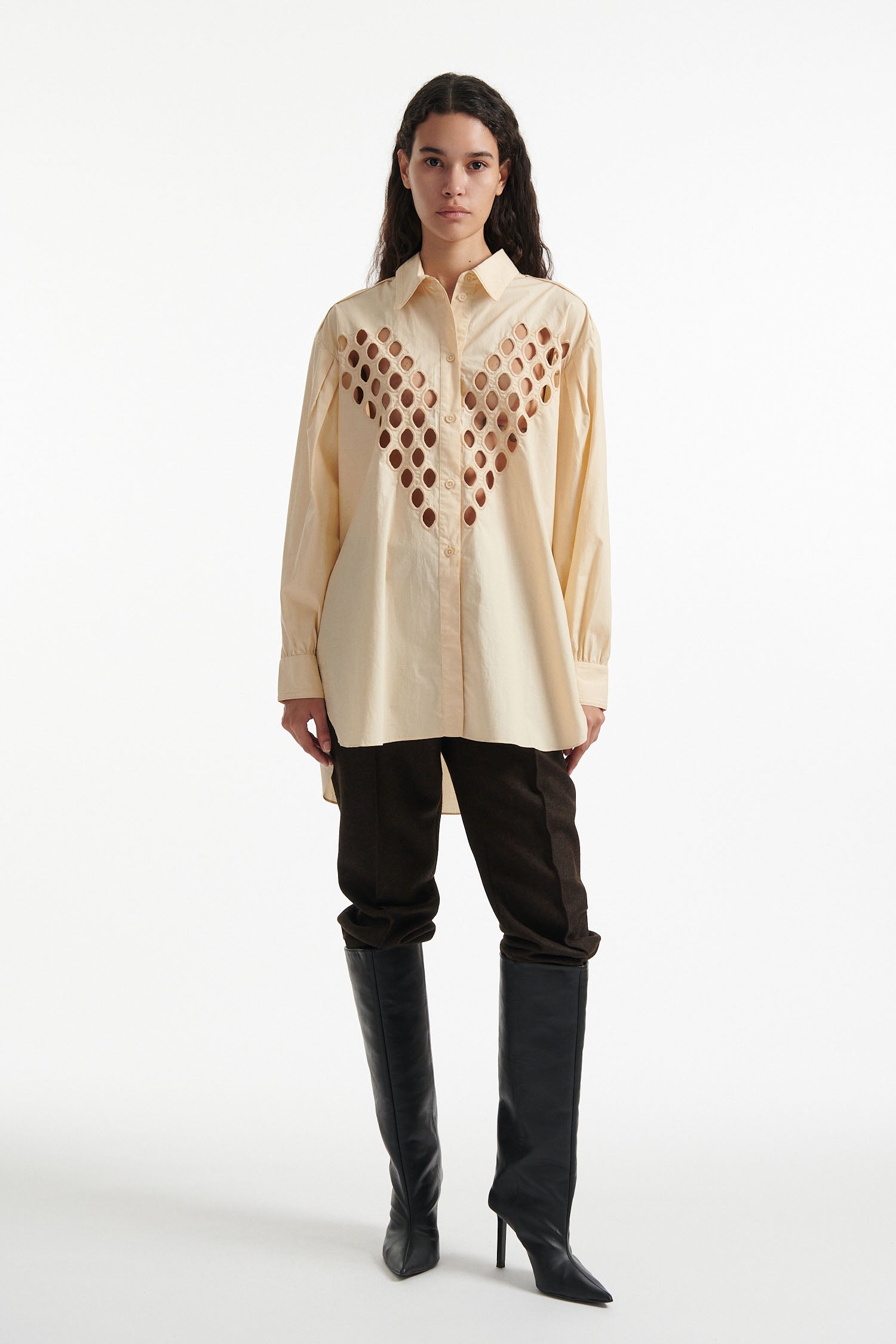 Female model wears off-white cotton shirt with see-through cutouts at chest and back. Model also wears brown wool pants and black high heels on white background.