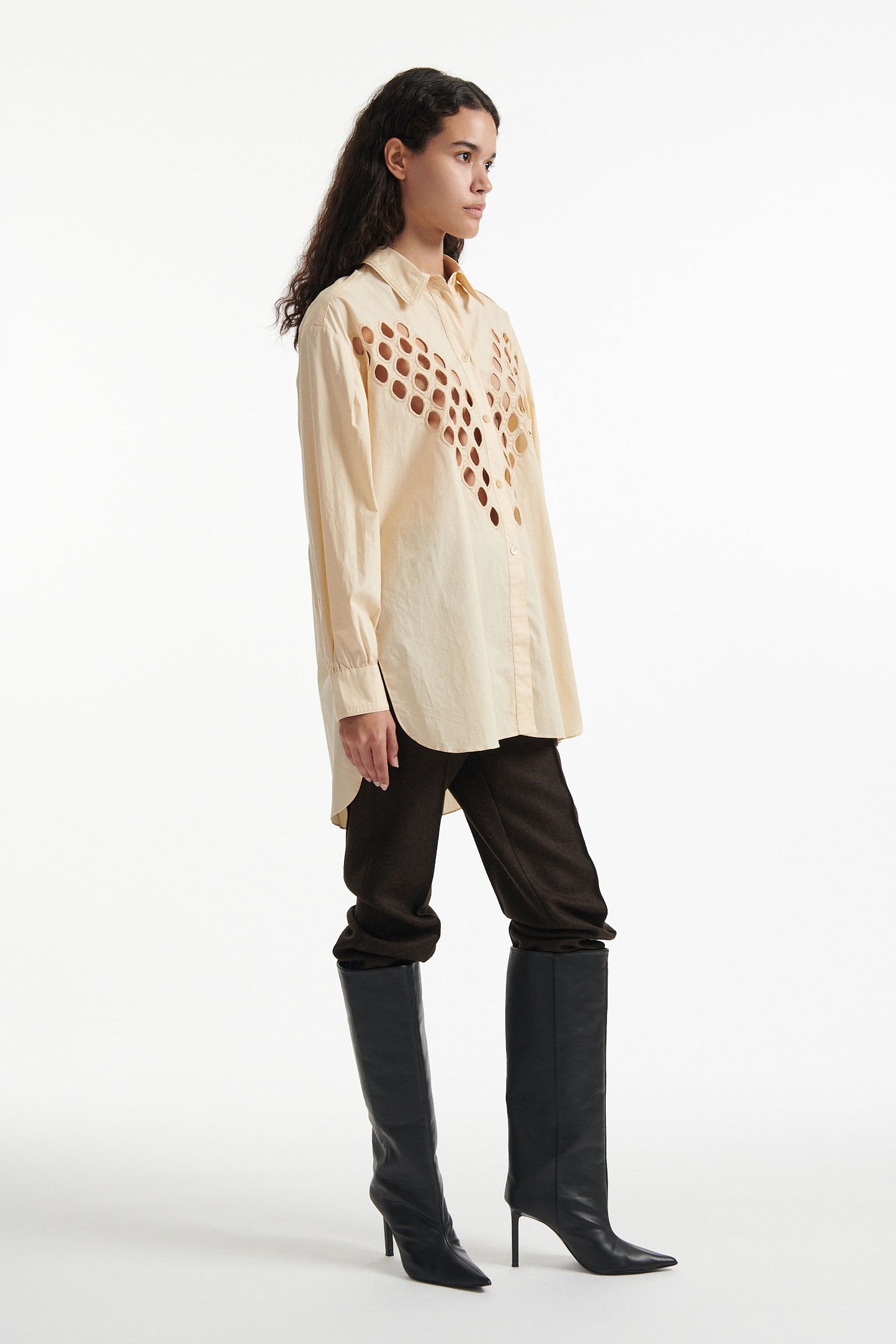 Female model wears off-white cotton shirt with see-through cutouts at chest and back. Model also wears brown wool pants and black high heels on white background.