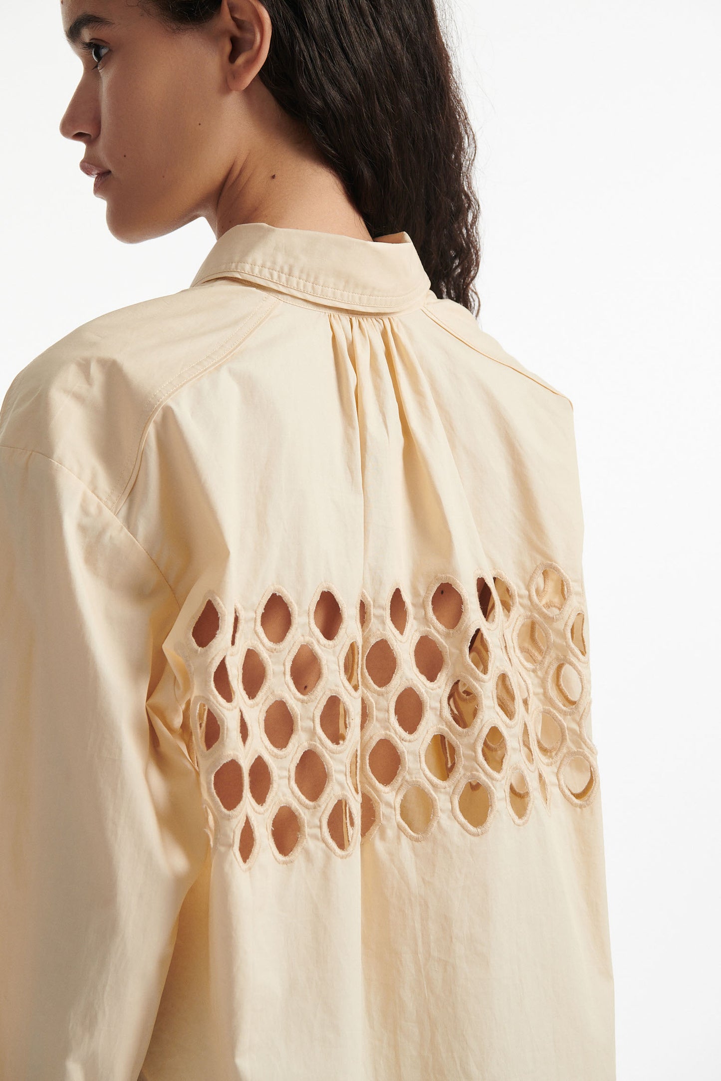 Female model wears off-white cotton shirt with see-through cutouts at chest and back. Model also wears brown wool pants and black high heels on white background.