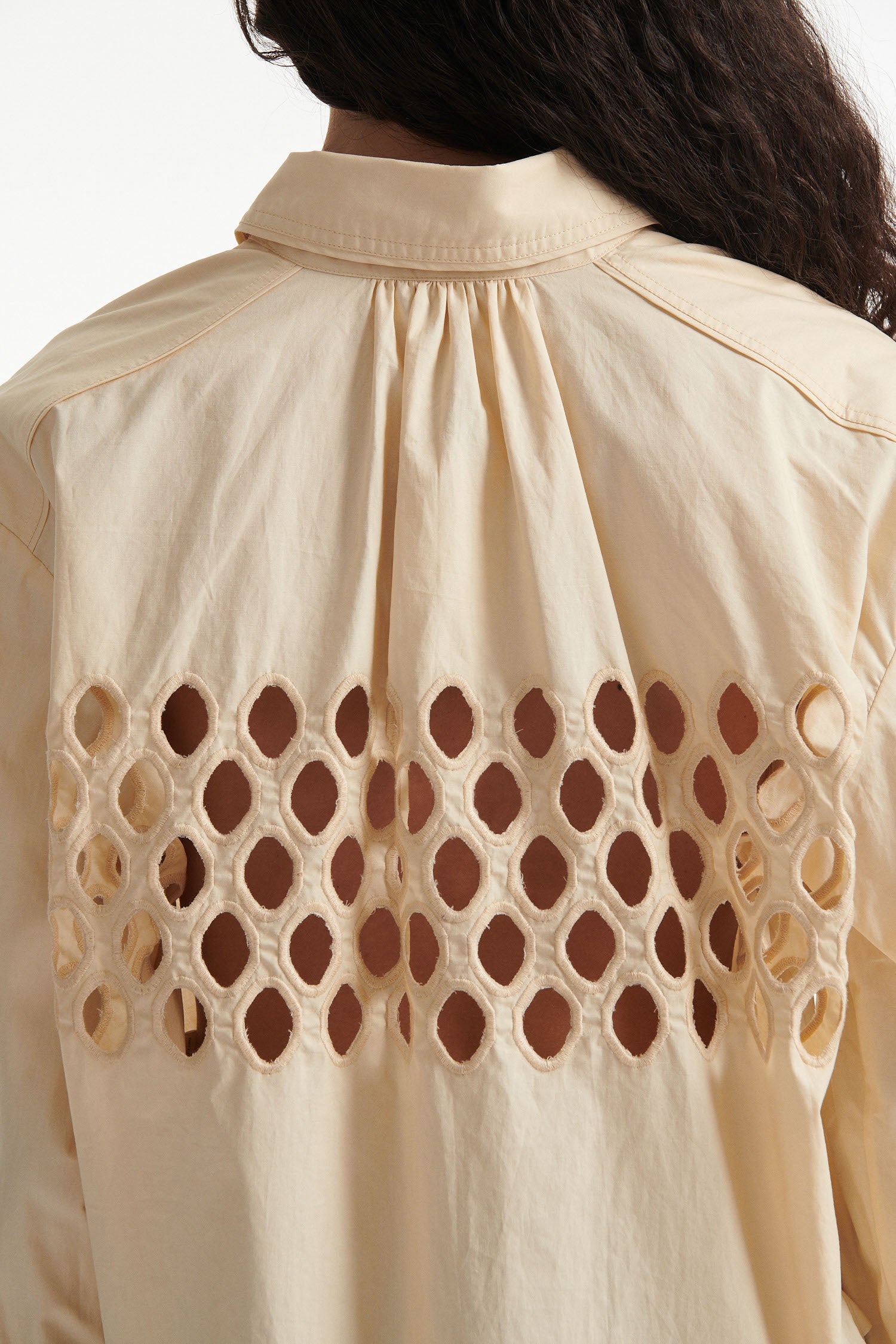 Female model wears off-white cotton shirt with see-through cutouts at chest and back. Model also wears brown wool pants and black high heels on white background.