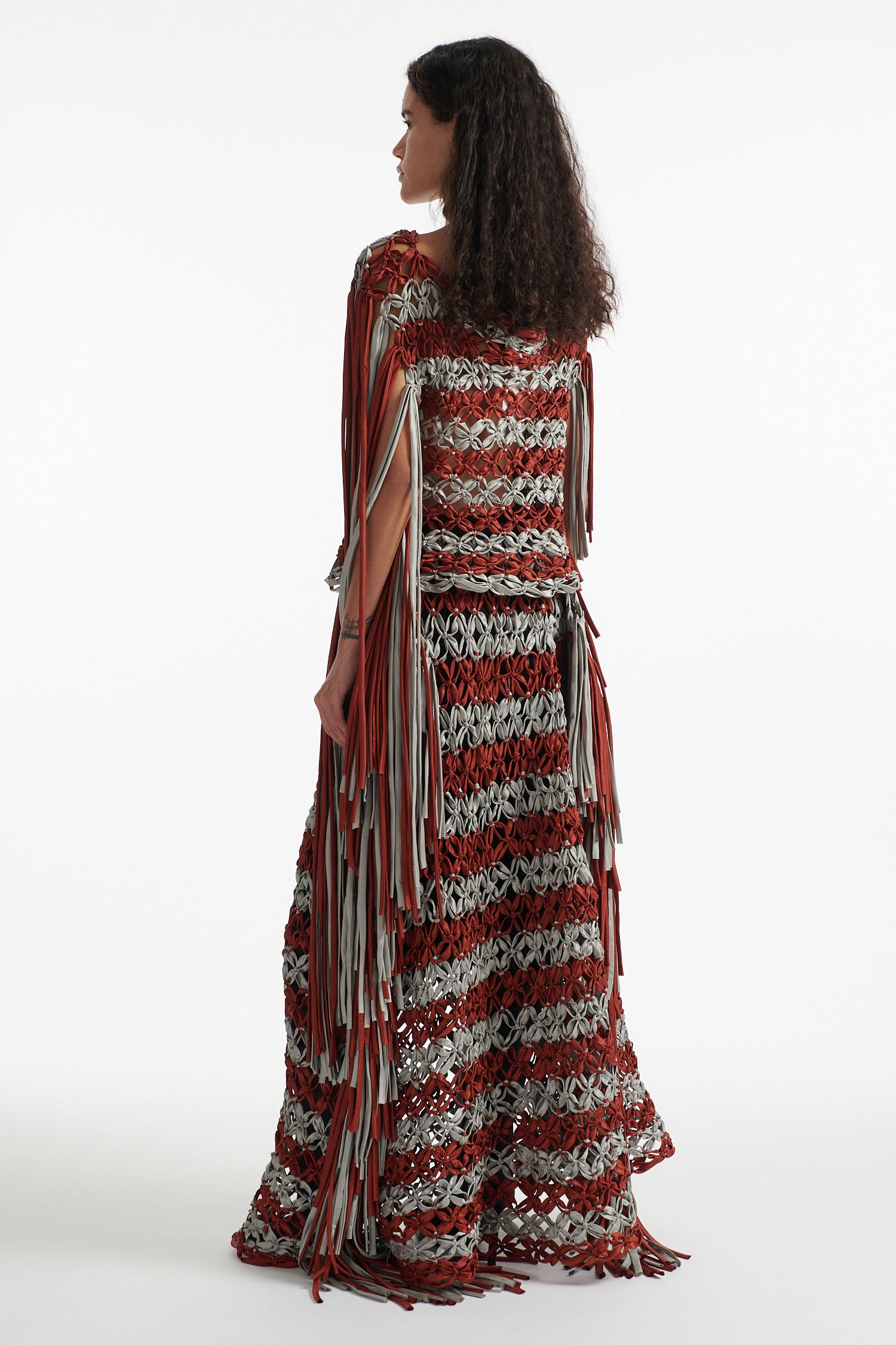 Female model wears knotted macrame set in burnt orange and sky grey. Both the top and skirt have asymmetric fringe hanging down either side. Model is on a white background.