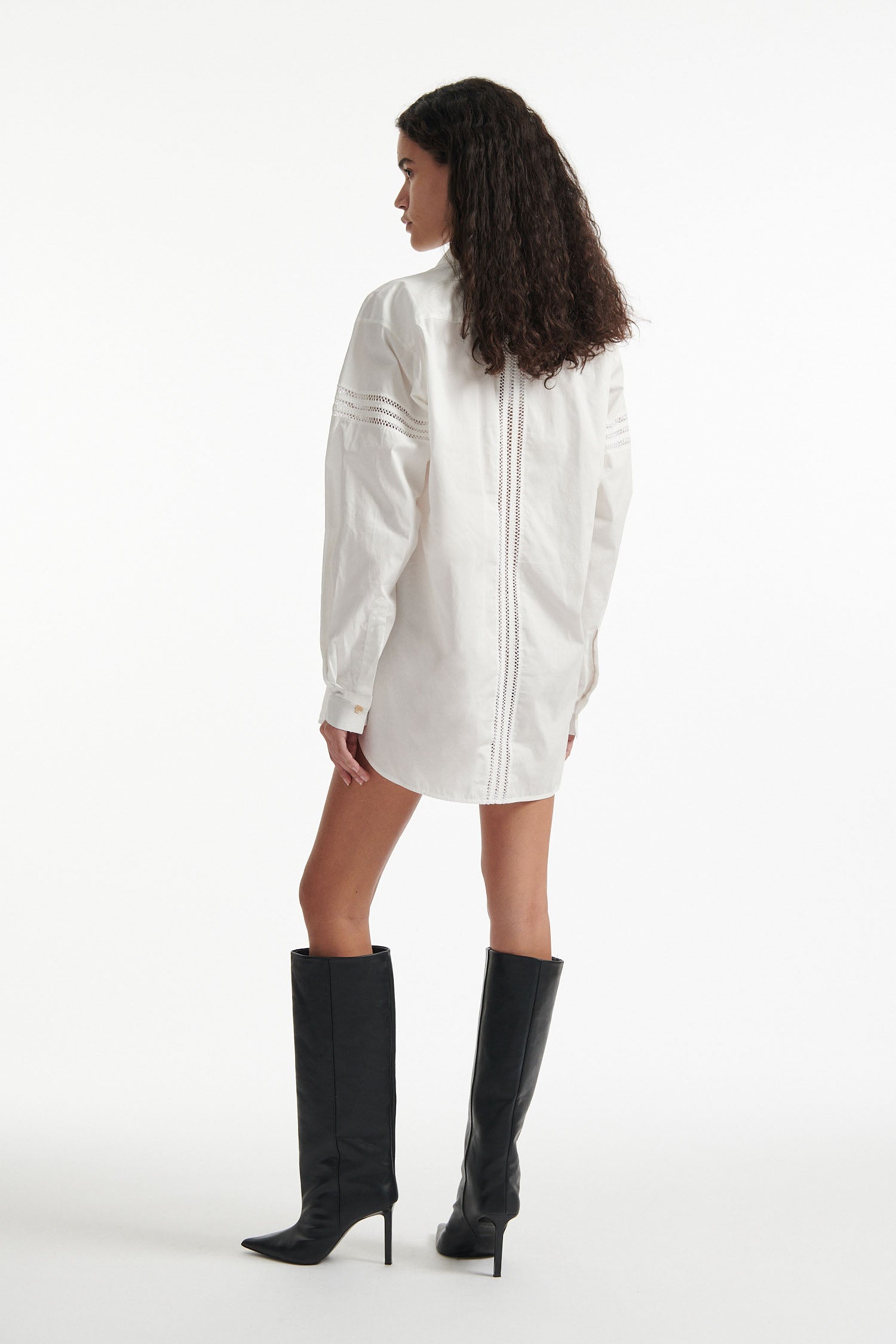 Female model wears white poplin shirt with exposed stitches and black high heel boots on white background.