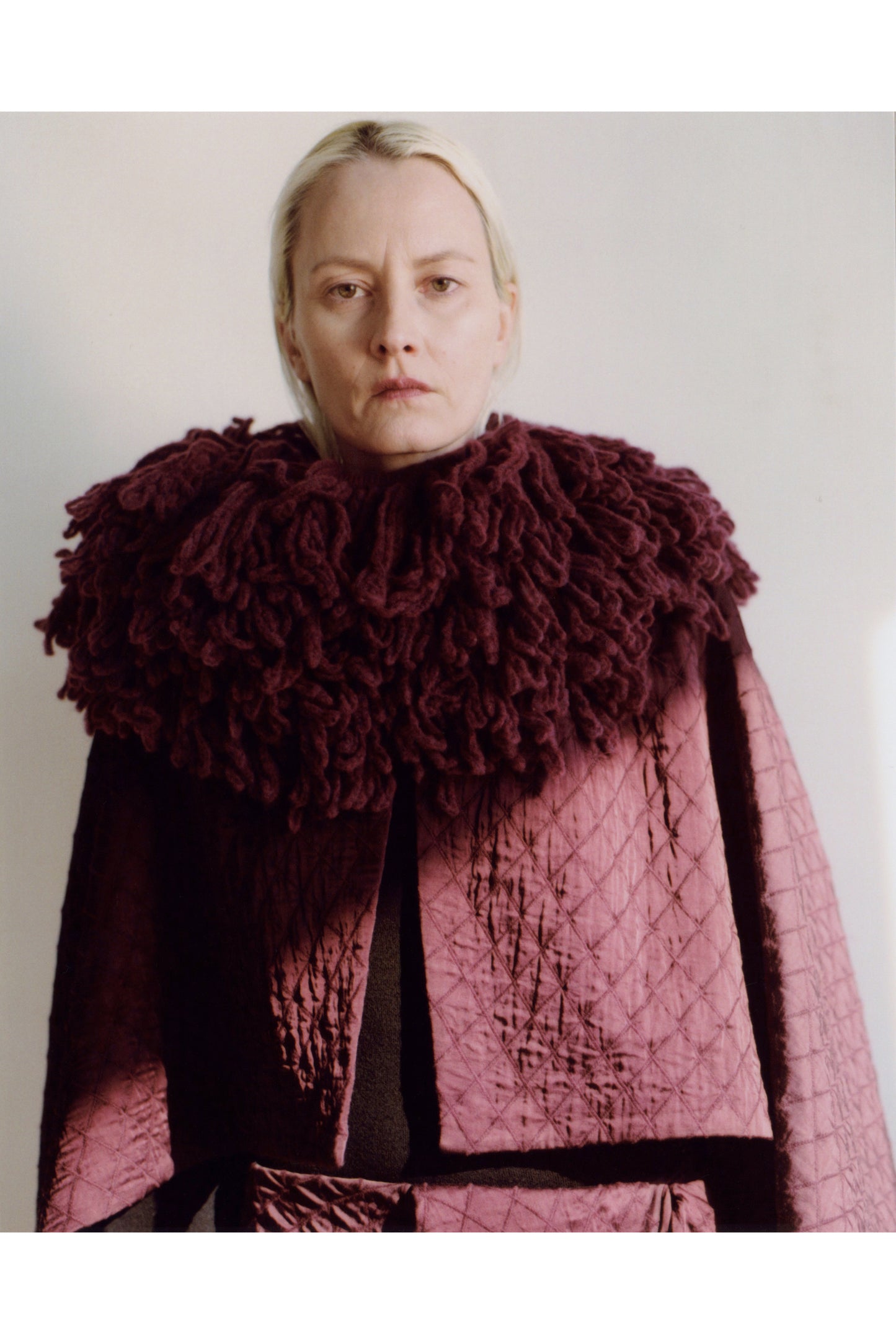 Person wearing a maroon cape with a ruffled collar against a plain background