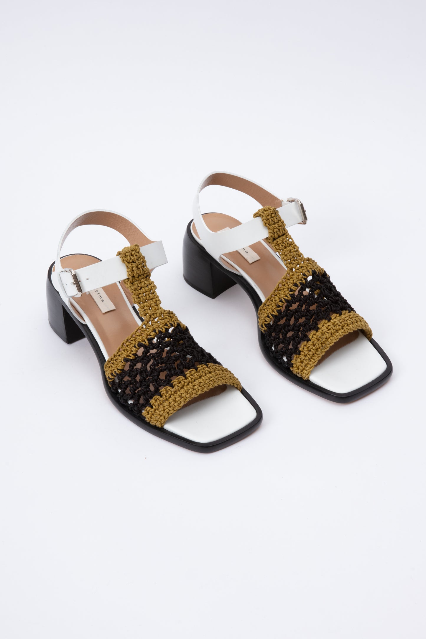 Open toe sandals with black leather base and white leather ankle straps. The block heel is slightly rounded and thick. Connected to the ankle strap is a toe strap made from hand-knit crochet in a cumin yellow and black.