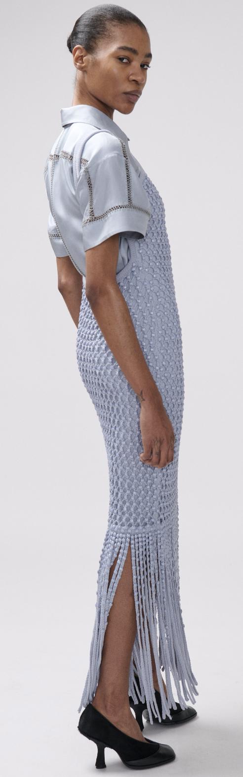 A model wearing a light blue crystal mesh gown with fringe detailing on the hem. Standing against a plain light gray background.