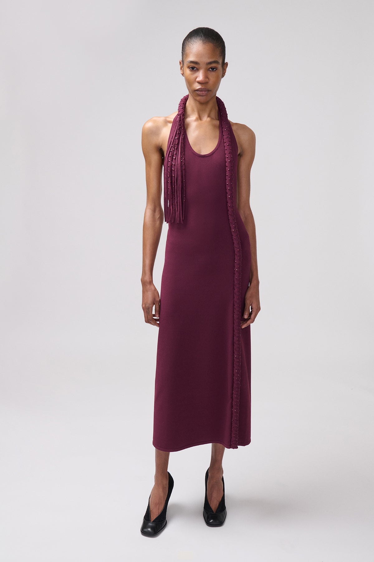 A model wearing a sleeveless, midi-length plum dress with a draped scarf detail with fringe.