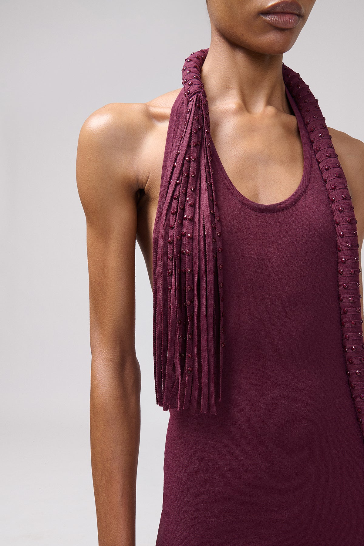 A close-up of a model wearing a deep burgundy halter-neck top with a matching fringed scarf draped around the neck, set against a plain light gray studio background.