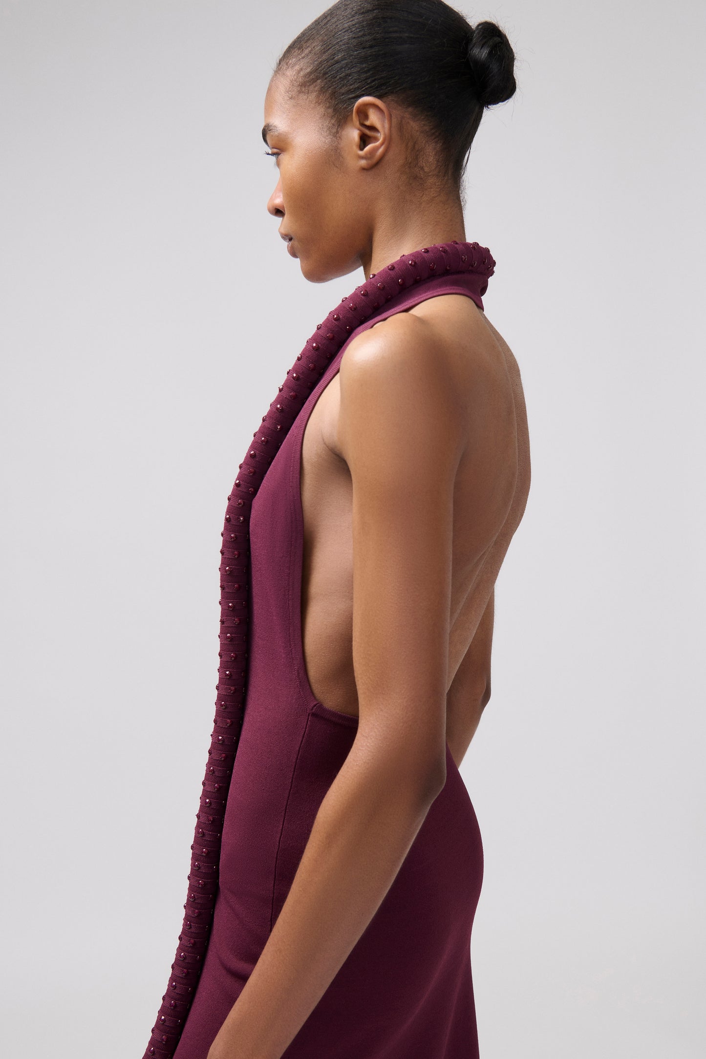 A model shown in profile wearing a sleeveless, deep plum-purple dress with a textured, ribbed knit collar and a low open back, set against a plain light gray studio background.