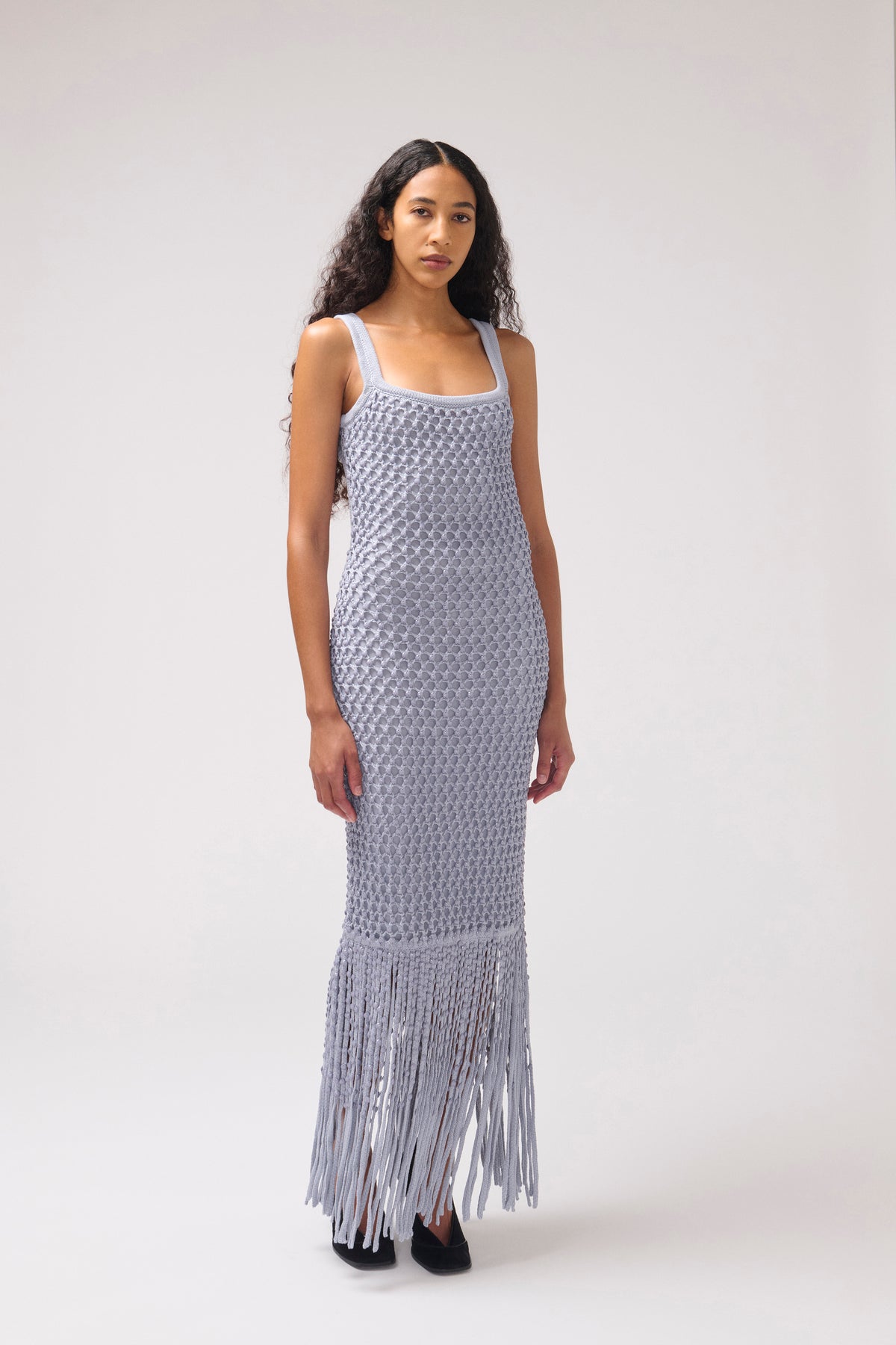 A model wearing a light blue-gray sleeveless crochet maxi dress with a fringed hem, standing against a neutral light gray studio background.