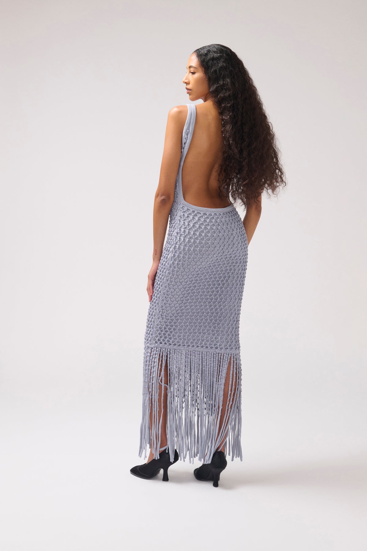 A woman stands with her back to the camera wearing a light blue open-back maxi dress with a textured crochet pattern and long fringe at the hem, paired with black heels, against a plain light gray studio background.