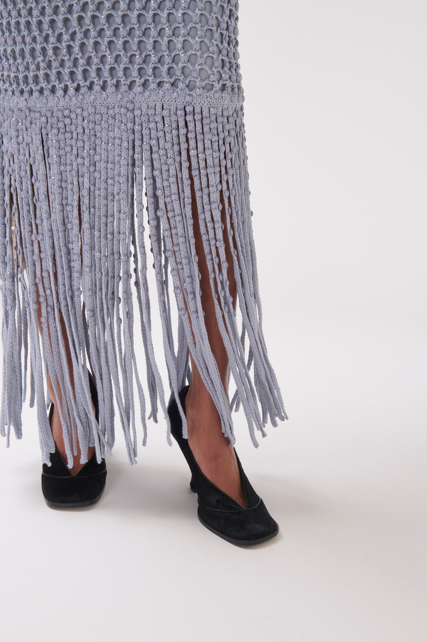 A close-up of a person wearing a light gray fringed knit skirt and black flat shoes against a plain white studio background.