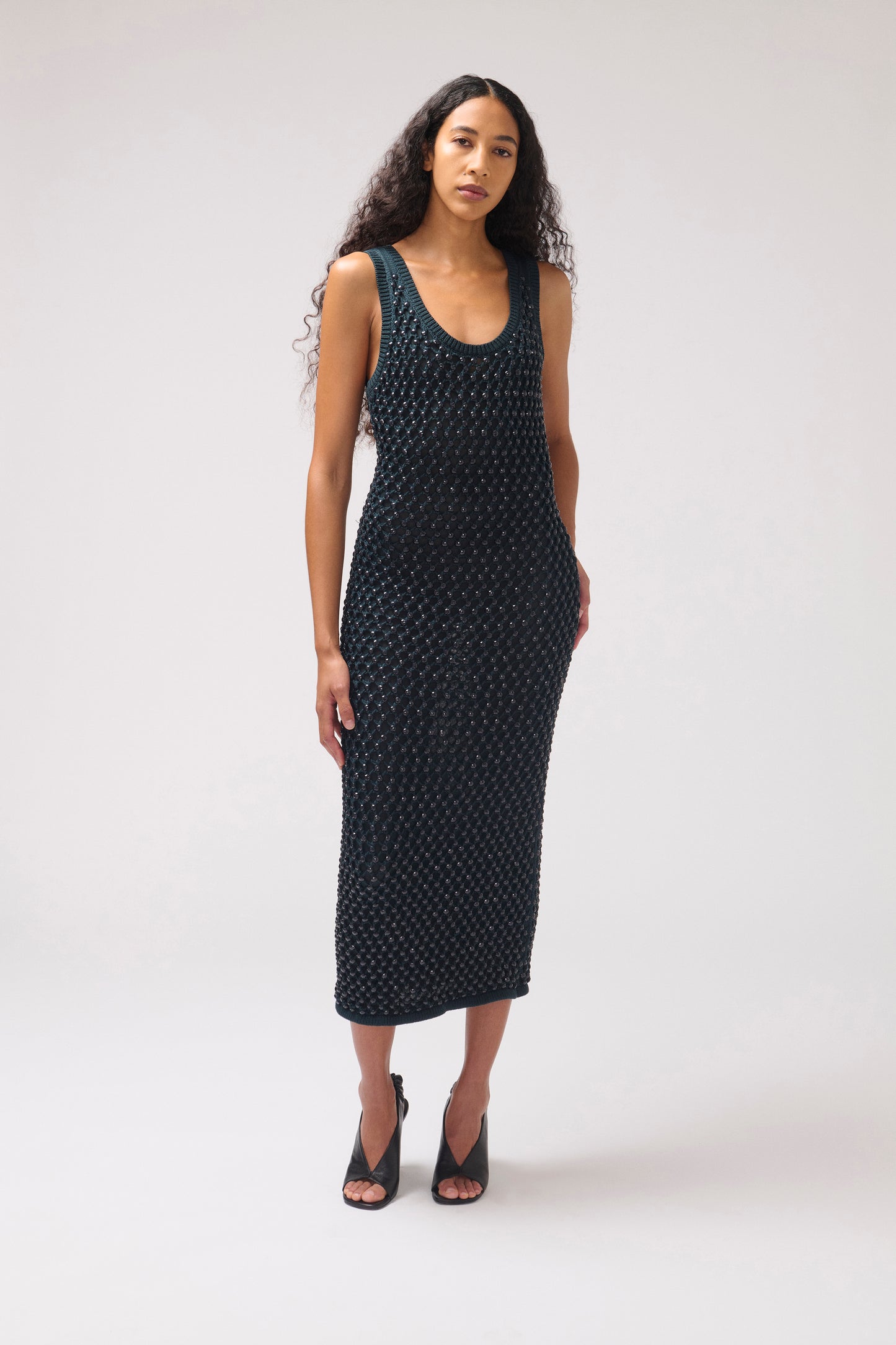 A woman modeling a sleeveless, knee-length black dress with a small white geometric pattern, paired with black strappy heels, against a light gray studio background.