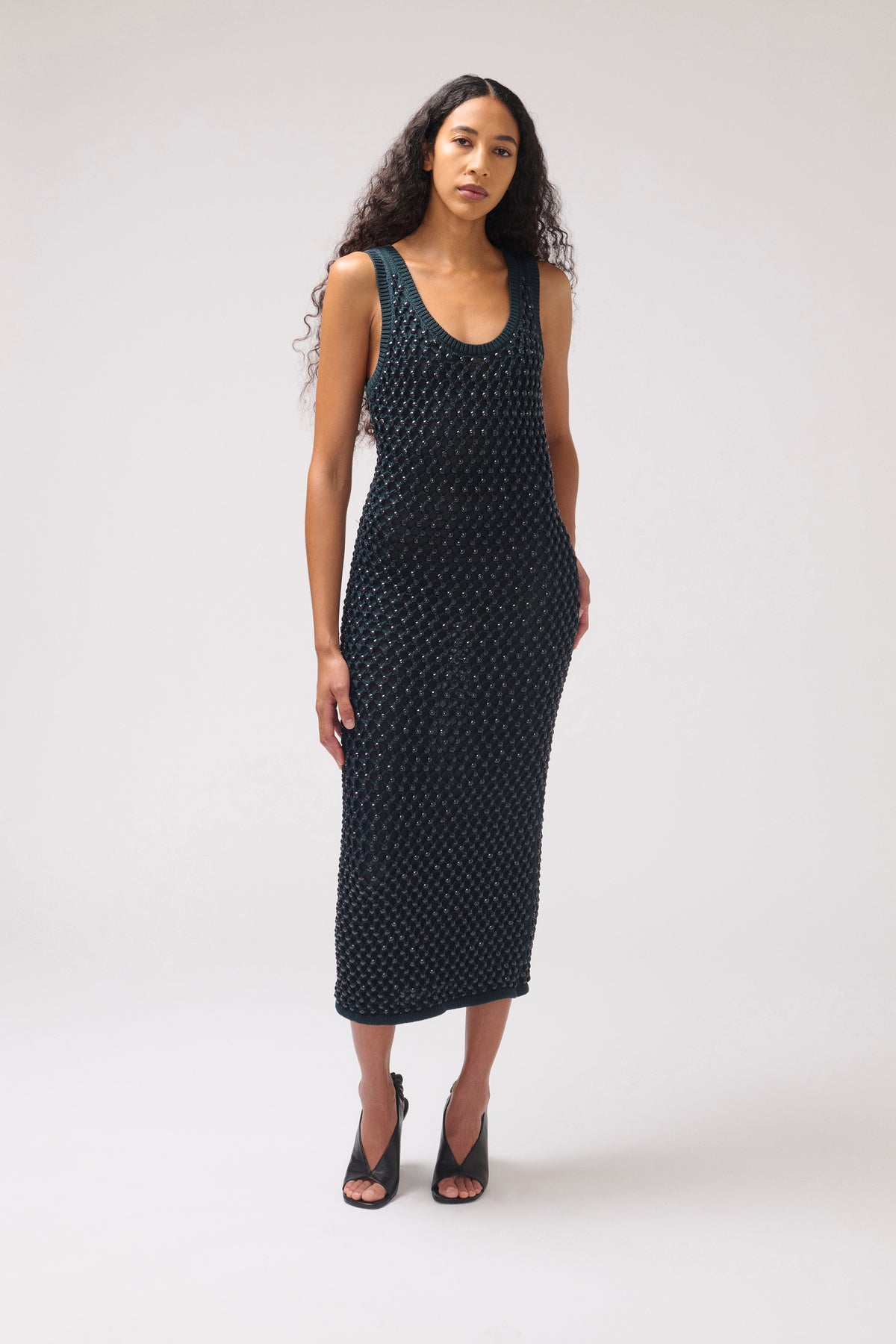 A woman modeling a sleeveless, knee-length black dress with a small white geometric pattern, paired with black strappy heels, against a light gray studio background.