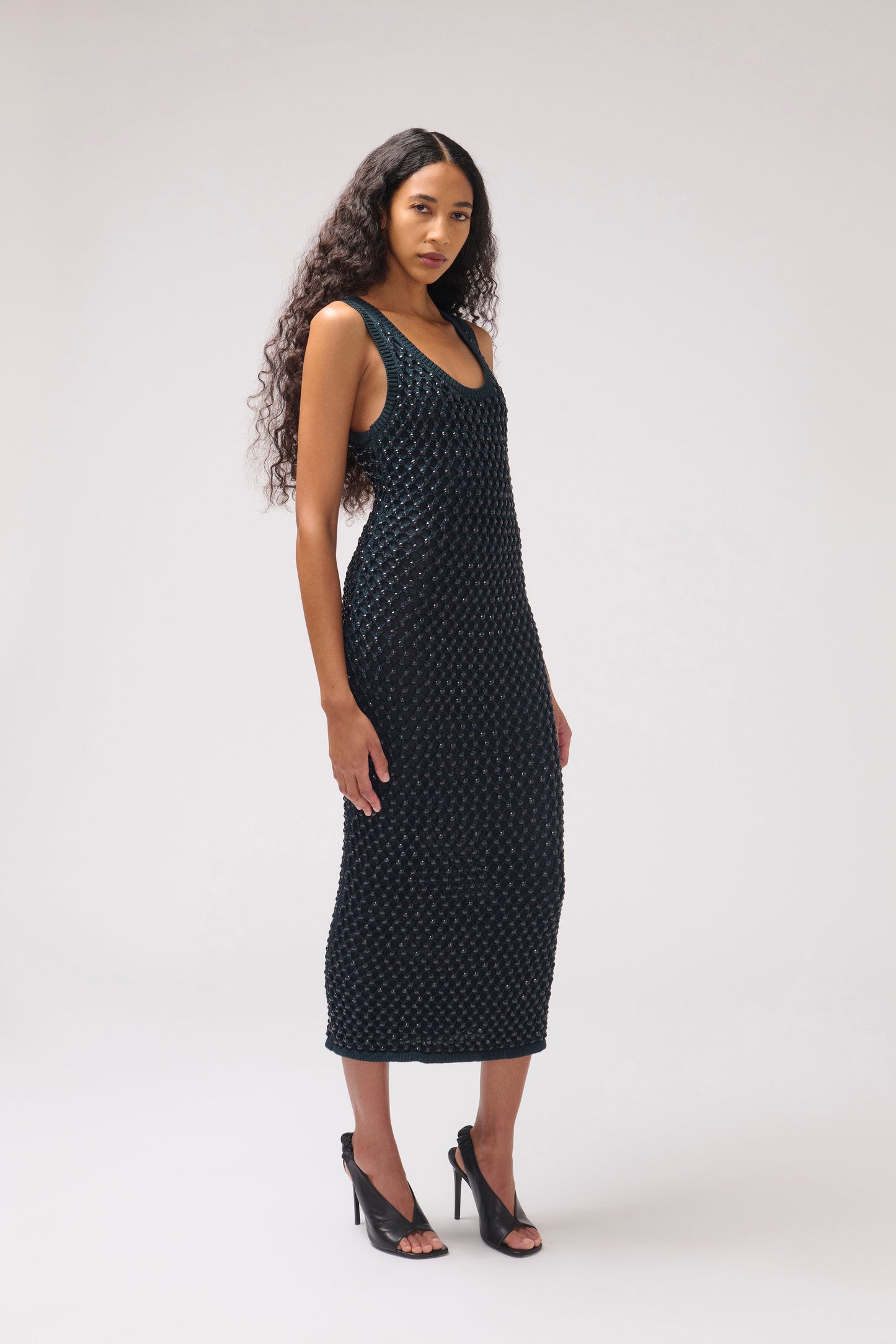 A woman modeling a sleeveless black midi dress with a small white geometric pattern, paired with black high-heeled sandals, against a light gray studio background.