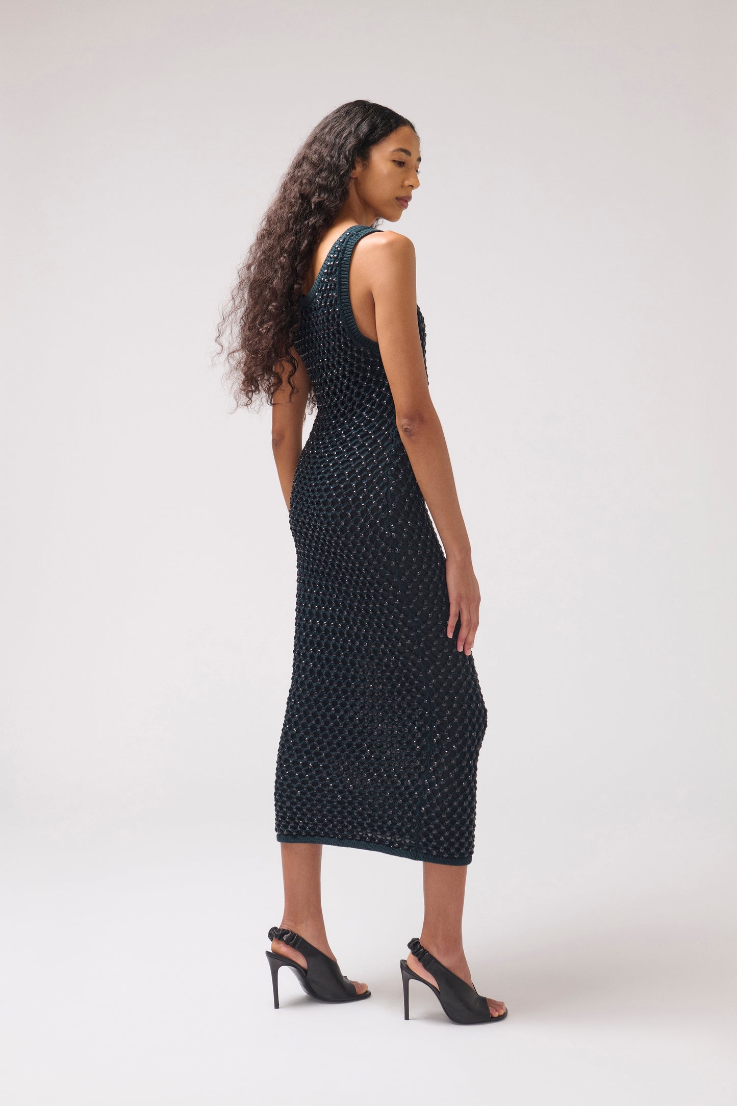 A woman modeling a sleeveless, floor-length black dress with a textured, dotted knit pattern, shown from a side-back angle against a plain light gray studio background.