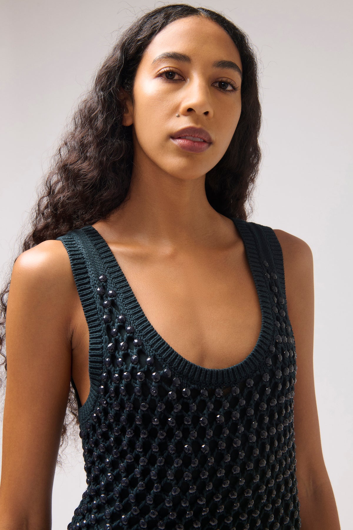 A woman wearing a dark green sleeveless knit top with a textured, beaded surface pattern, photographed against a neutral light gray background.