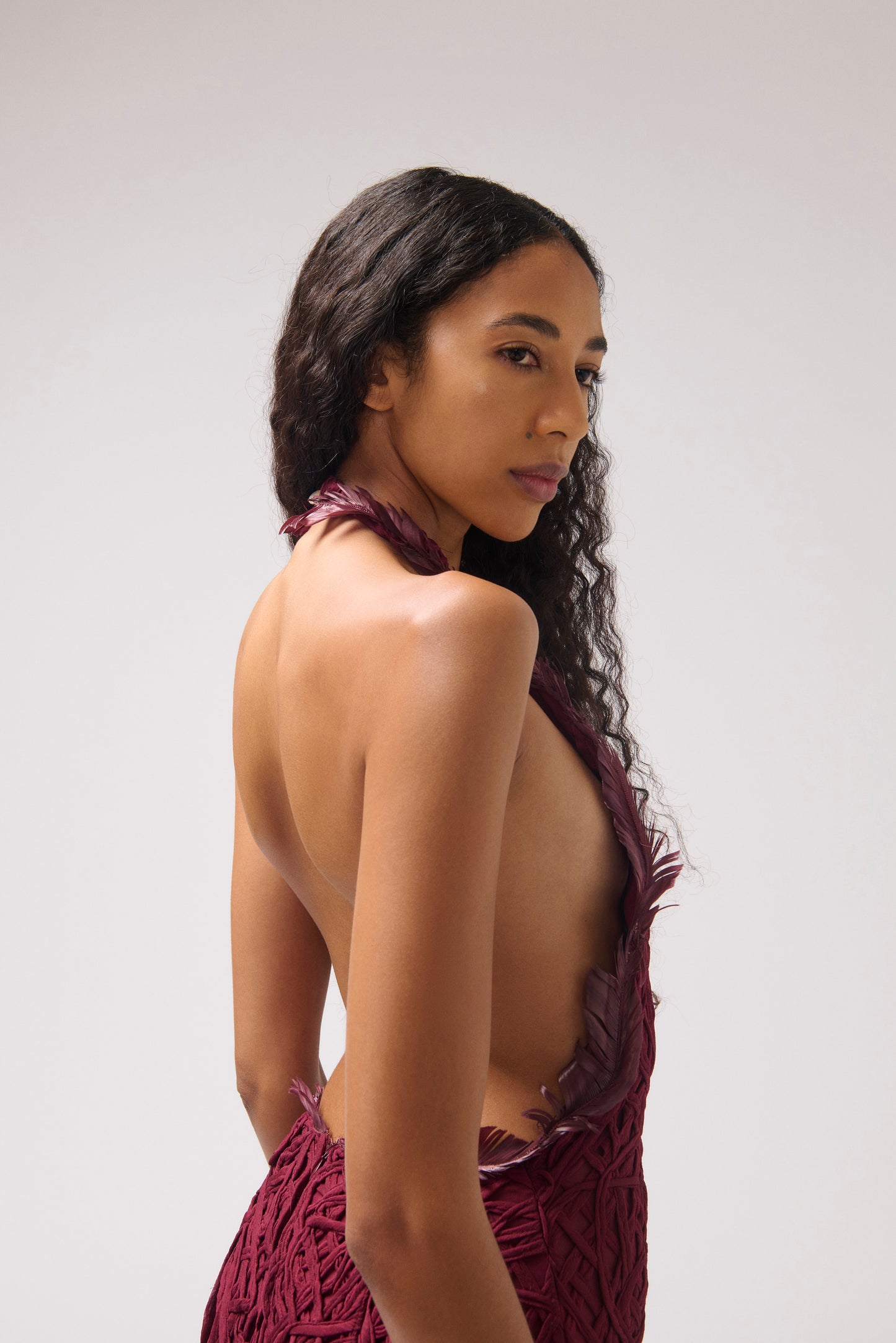A model shown from a side-back angle, wearing a textured halter dress in a deep burgundy color. The dress features burgundy feather details and a large open back.