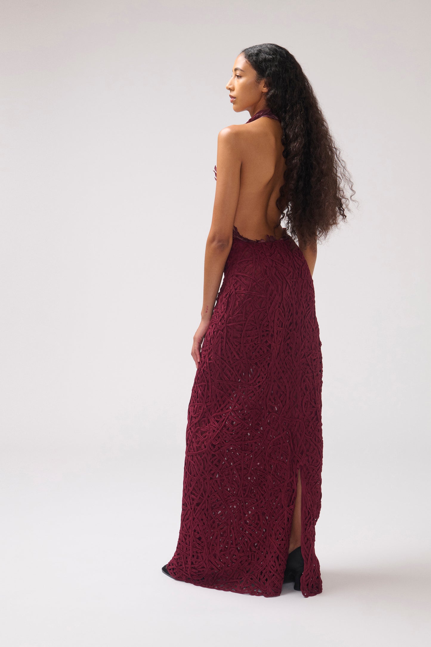A model in front of a white background shown in profile. The model is wearing a textured halter dress in a deep burgundy color. The dress features burgundy feather details and a large open back.