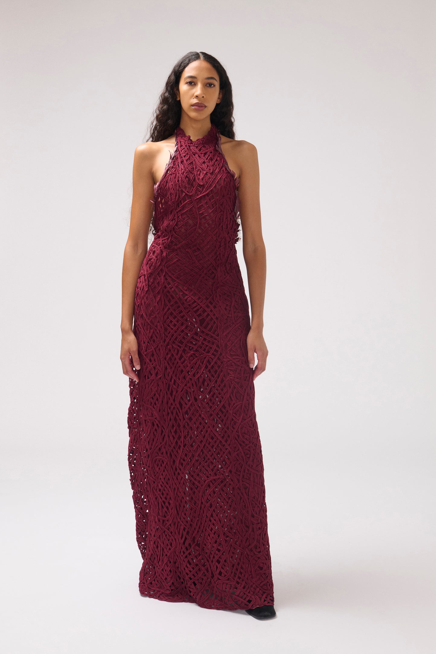 A model in front of a white background, wearing a textured halter dress in a deep burgundy color. The dress features burgundy feather details and a large open back.