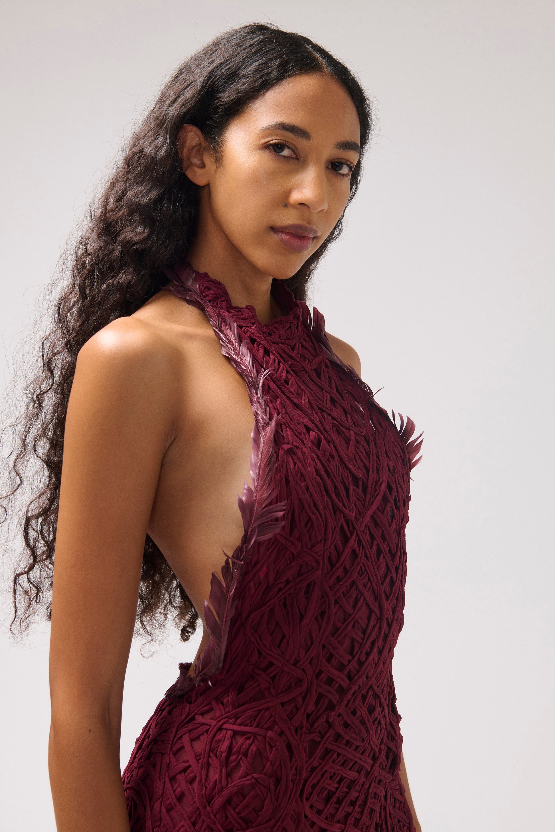 A model in front of a white background, wearing a textured halter dress in a deep burgundy color. The dress features burgundy feather details and a large open back. The image is cropped to the upper half of the dress.