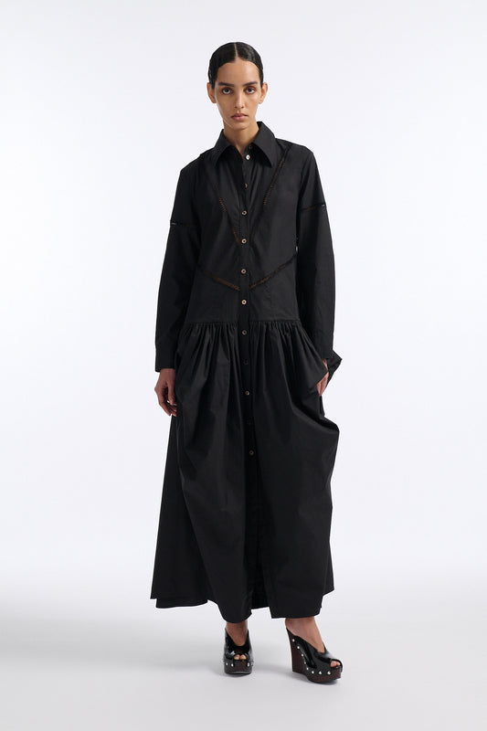 Female model with slicked back black hair in a bun wears black button down collared dress with diagonal hand stitched detailing along front, back and sleeves. The waist of the skirt is rouched to provide volume. The model also wears white palm cutout denim. 