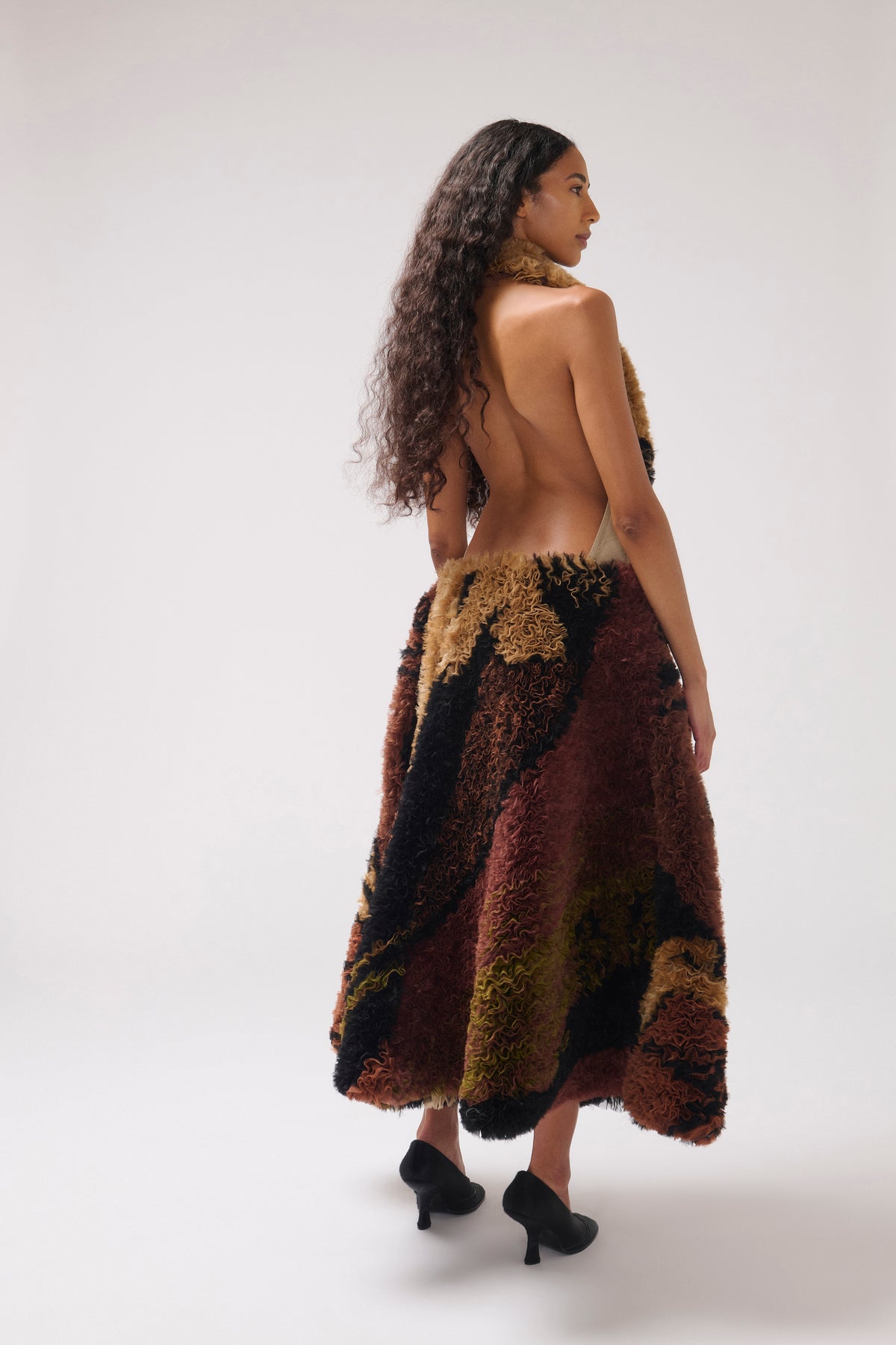 A model is shown from behind wearing a shaggy, textured halter maxi dress. The dress features an abstract pattern of rust red, olive green, black, and brown.