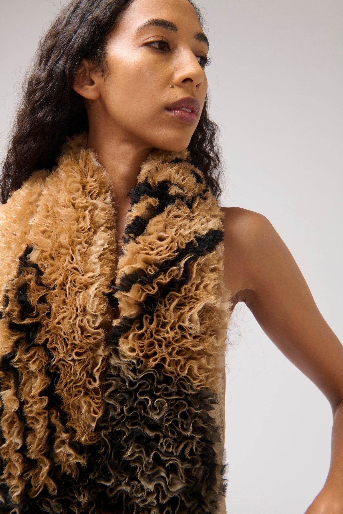 A close-up of a model wearing a chunky, curly shaggy halter dress in a brown and black pattern against a plain light gray background.