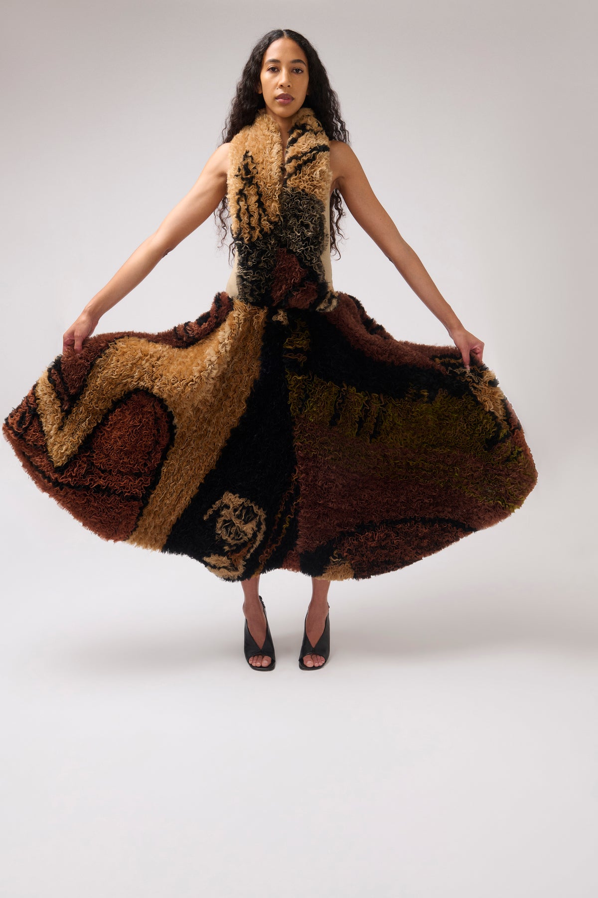 A model wearing a high-pile textured halter dress, holding out the wide skirt to display its volume. The dress features an abstract pattern of brown, tan, black, and olive green.