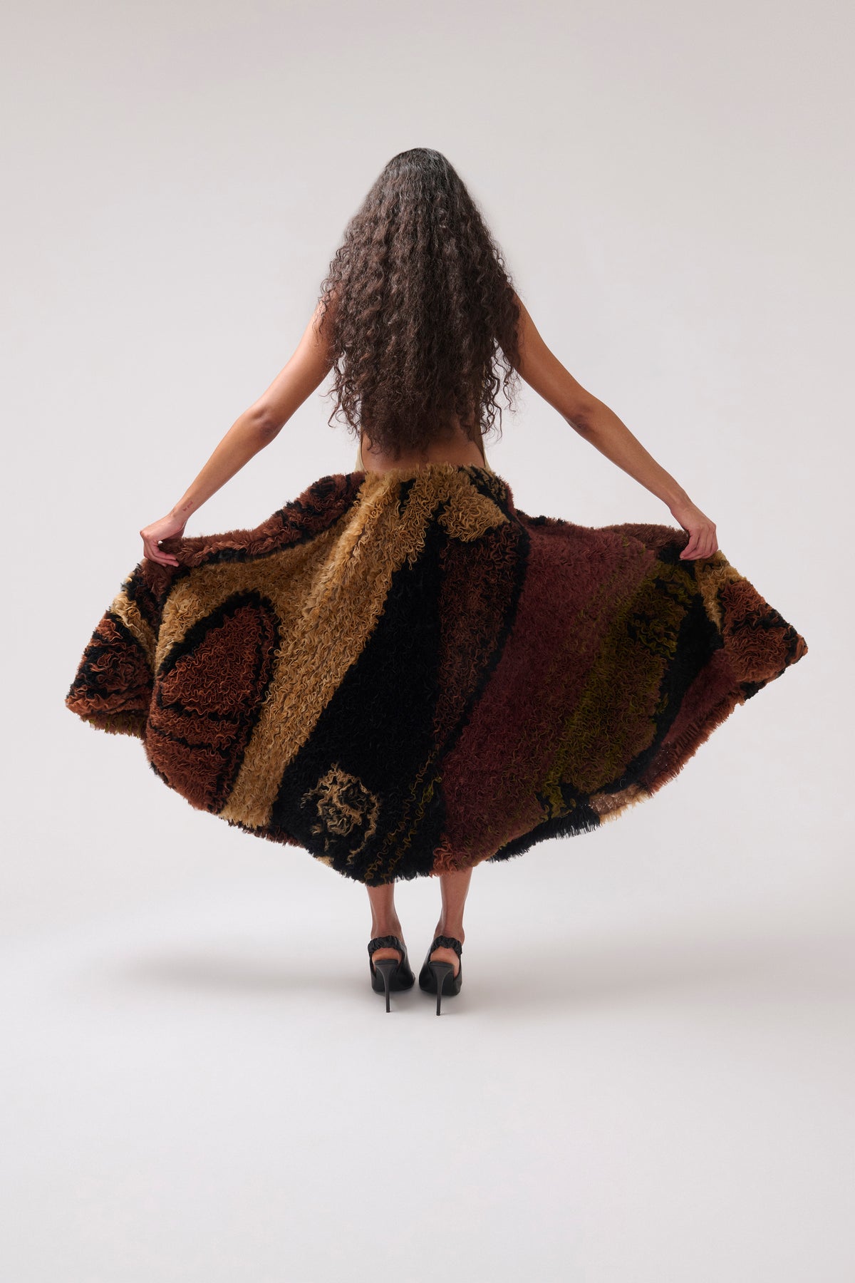 A woman viewed from behind wearing a voluminous shaggy halter dress made of mixed brown, black, tan, and burgundy textured fabric, holding the sides out to show its shape against a plain light gray studio background.