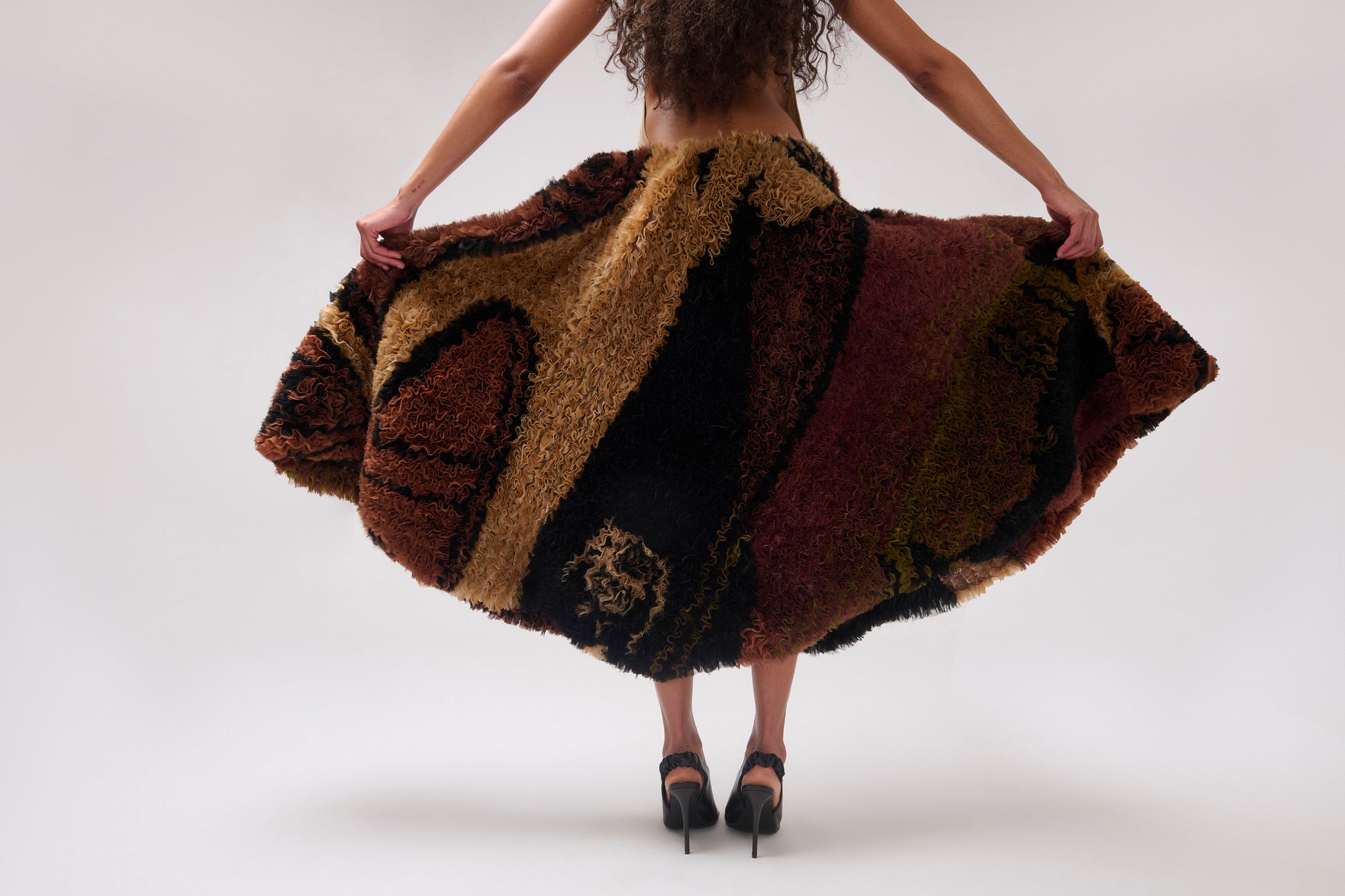 A woman viewed from behind wearing a voluminous printed organza skirt. holding the skirt out against a plain light gray studio background.