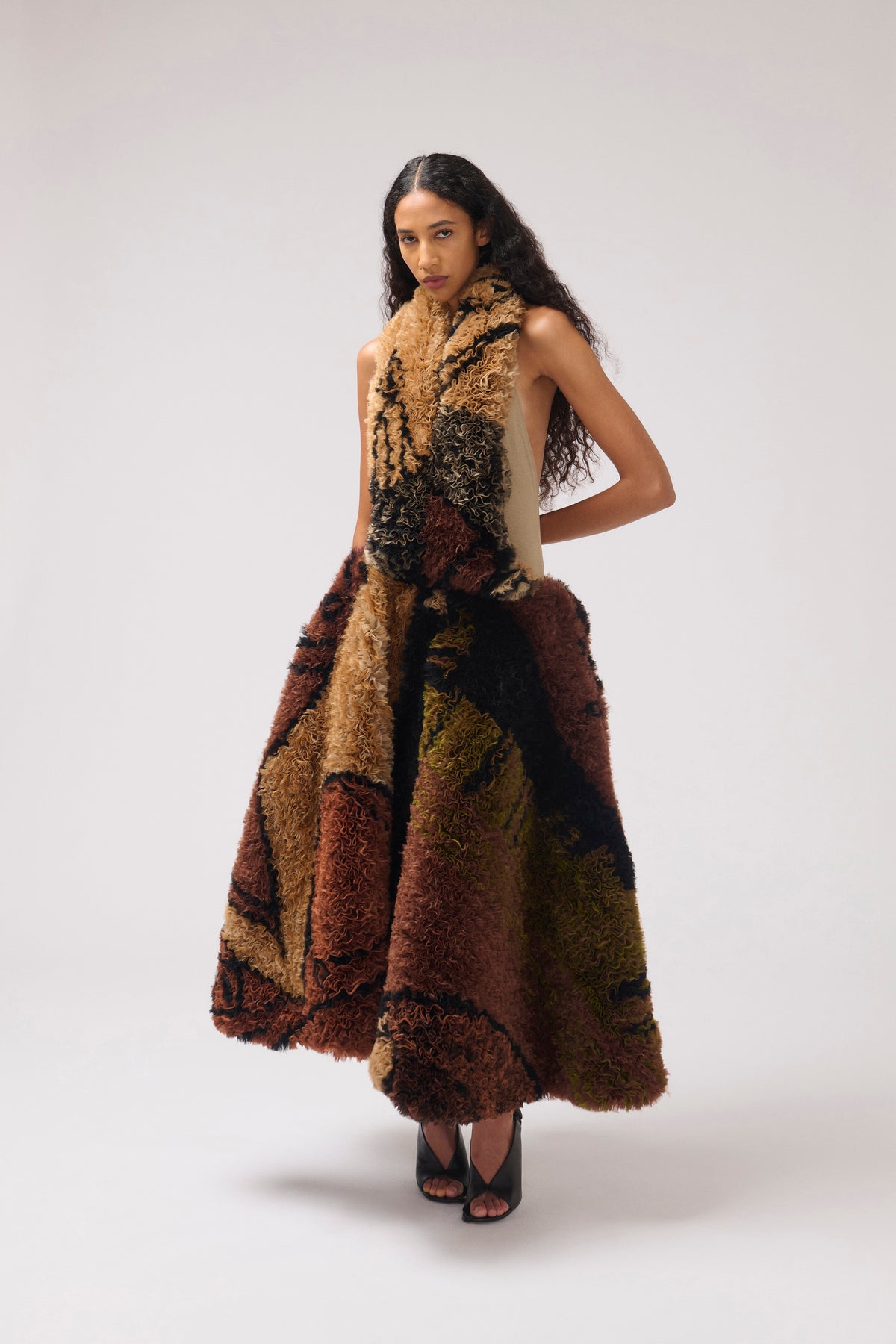 A model wearing a sleeveless, high-neck, voluminous shaggy dress with a patchwork pattern of brown, beige, and black, photographed against a neutral light gray studio background.